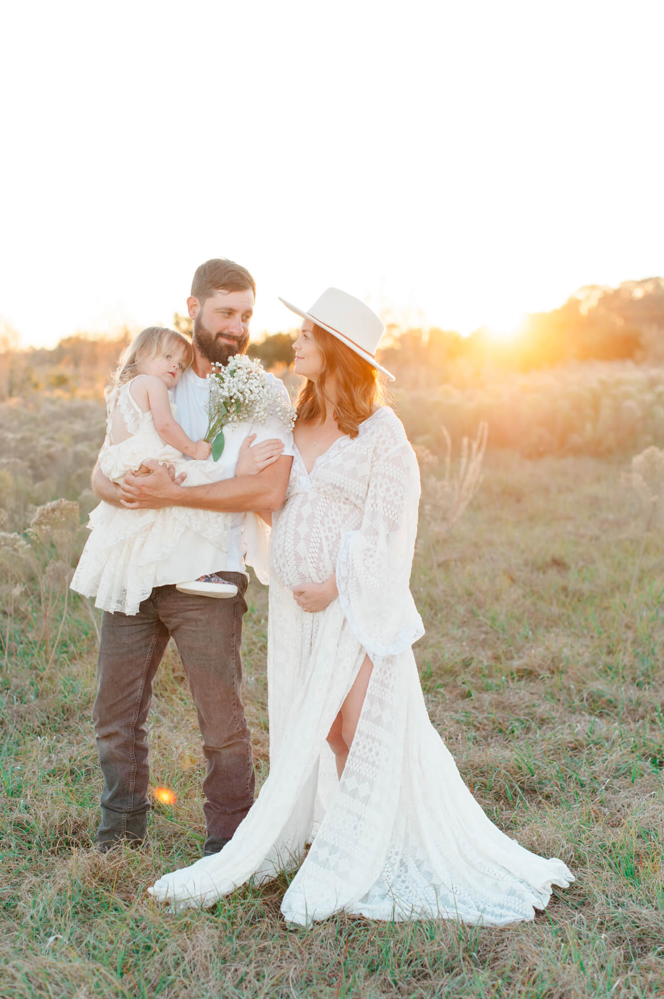 Family of soon to be four embrace while standing in a field with a beautiful sunset glow behind them 