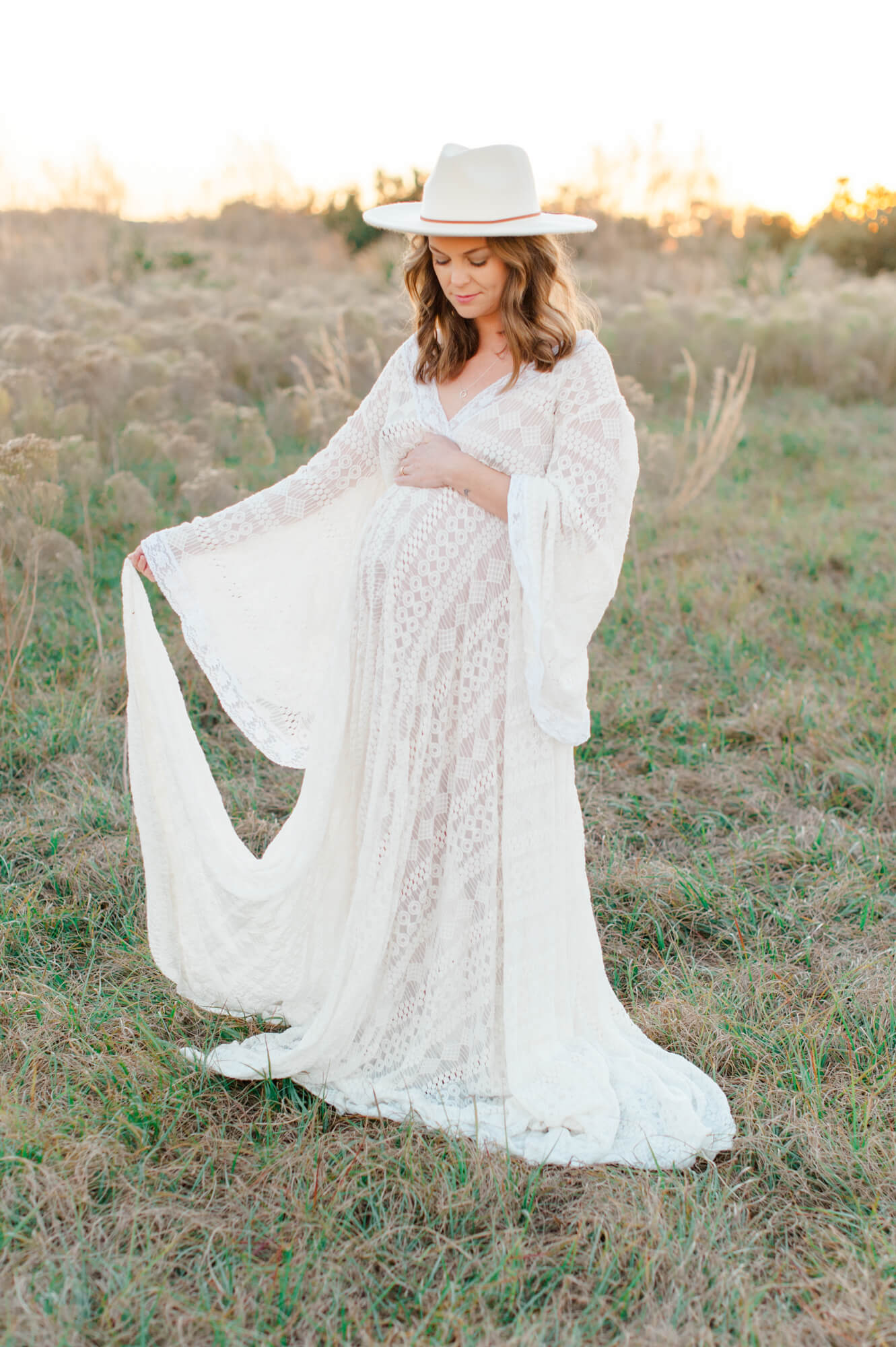 Stunning expectant mother stands waving her white lace gown in a tall grass field during golden hour Space Coast Doulas