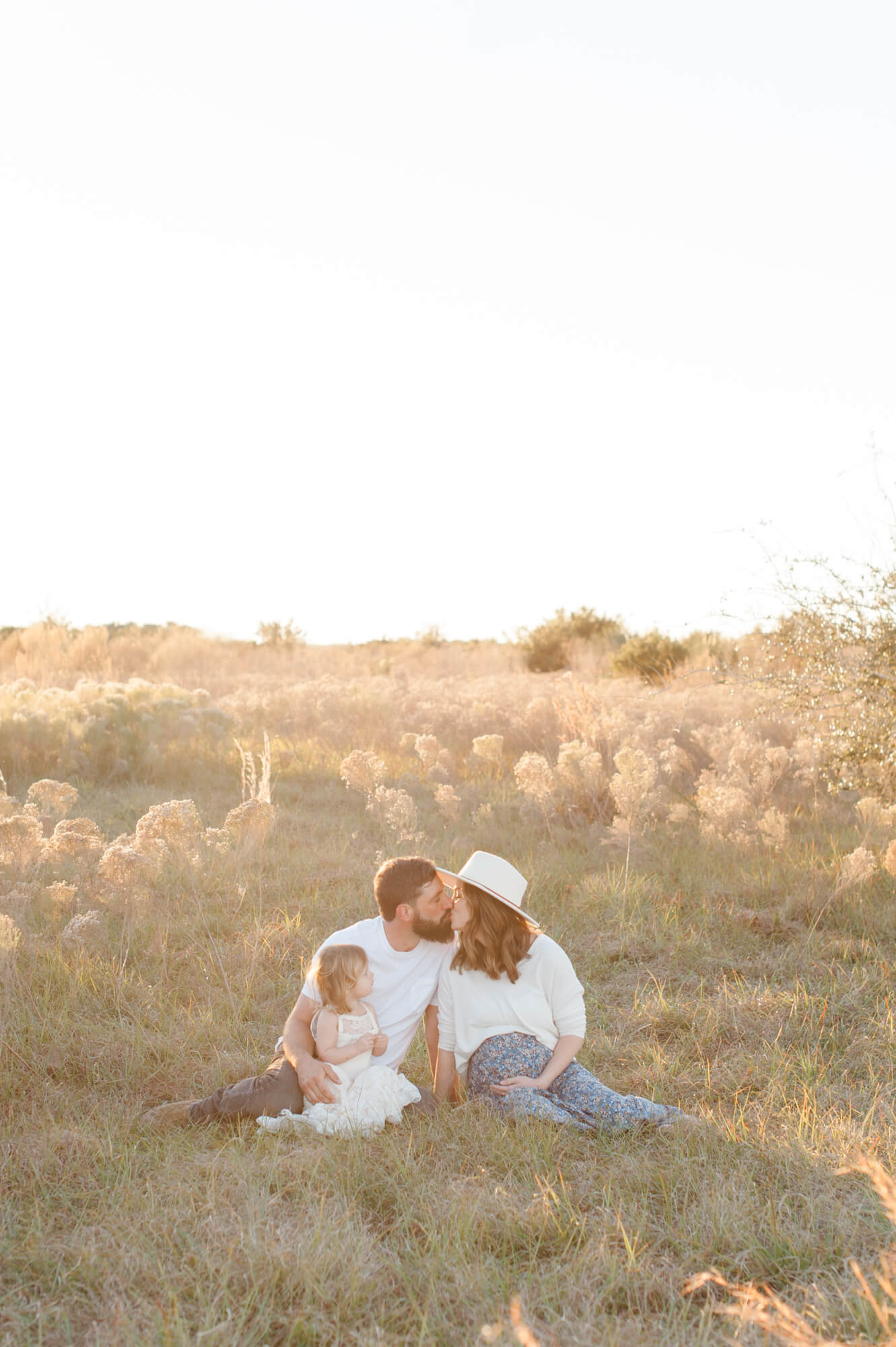 Expecting parents share a kiss while sitting in a tall grass field with their young toddler daughter at sunset.
