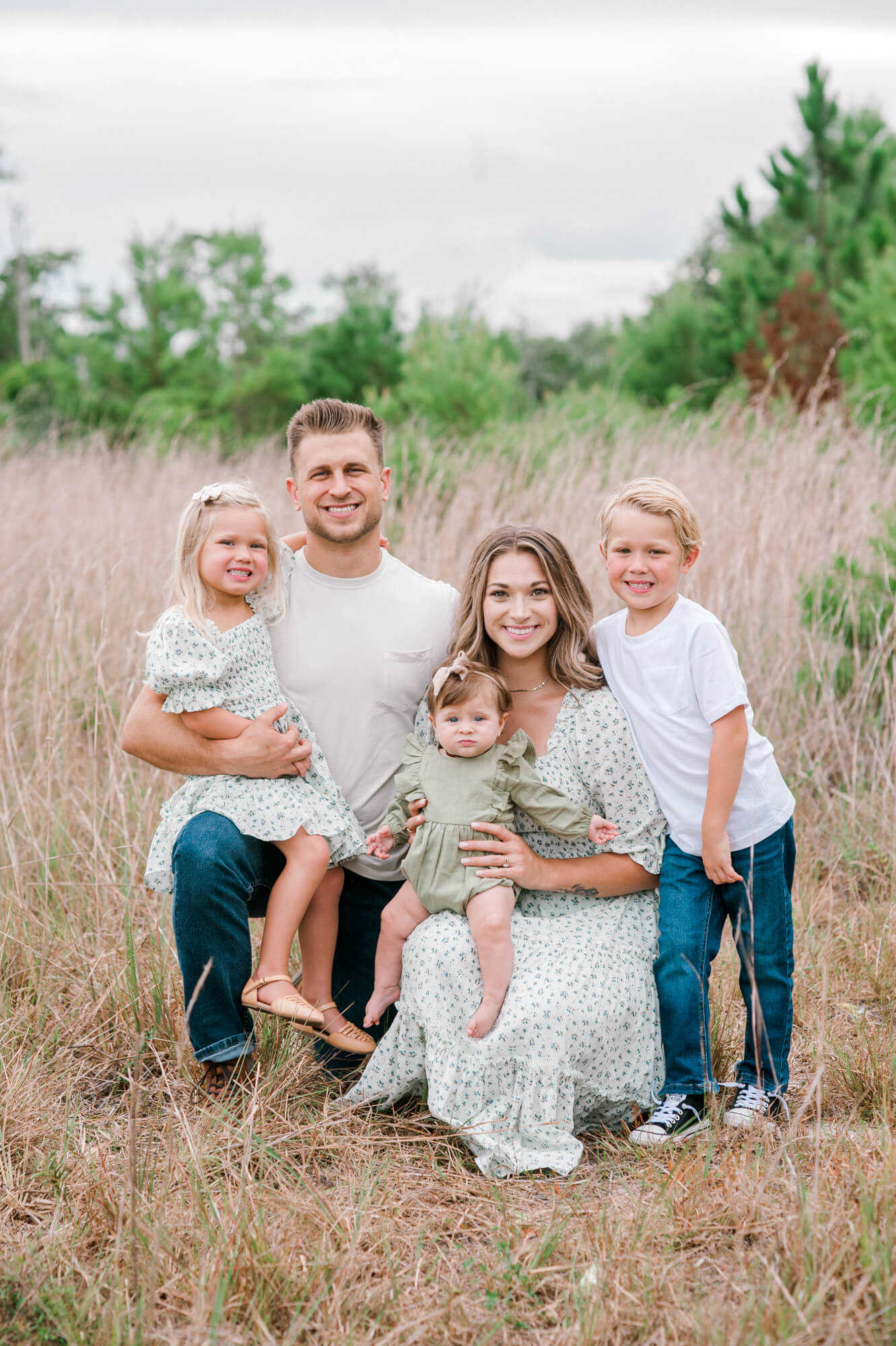 Family of five smiling and looking at the camera in the middle of a tall grass field. 