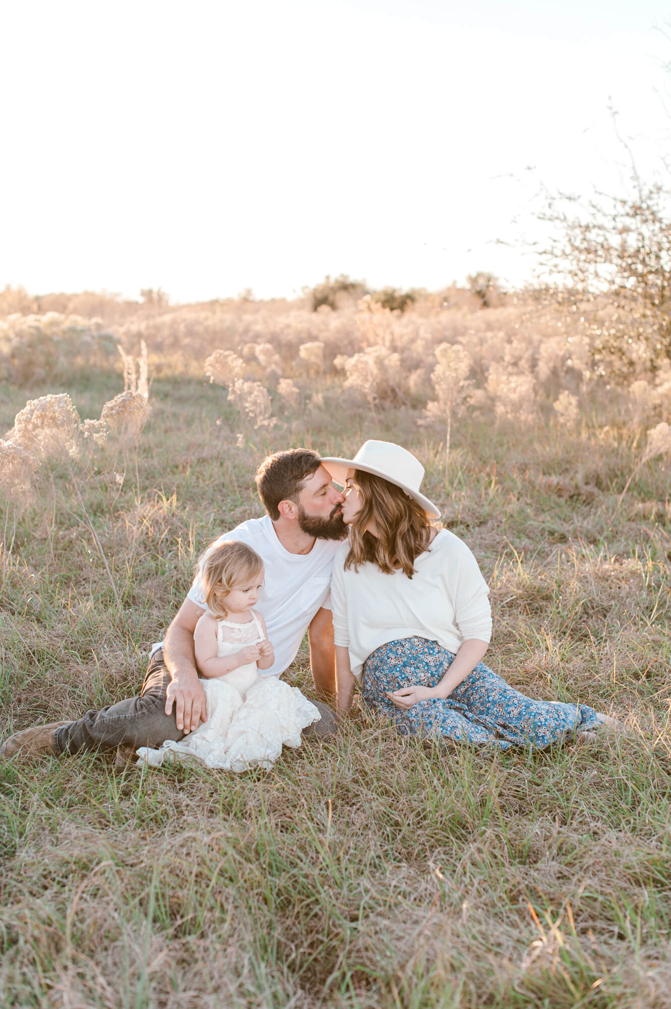Mom and dad to be sit in a field with their daughter kissing. 
