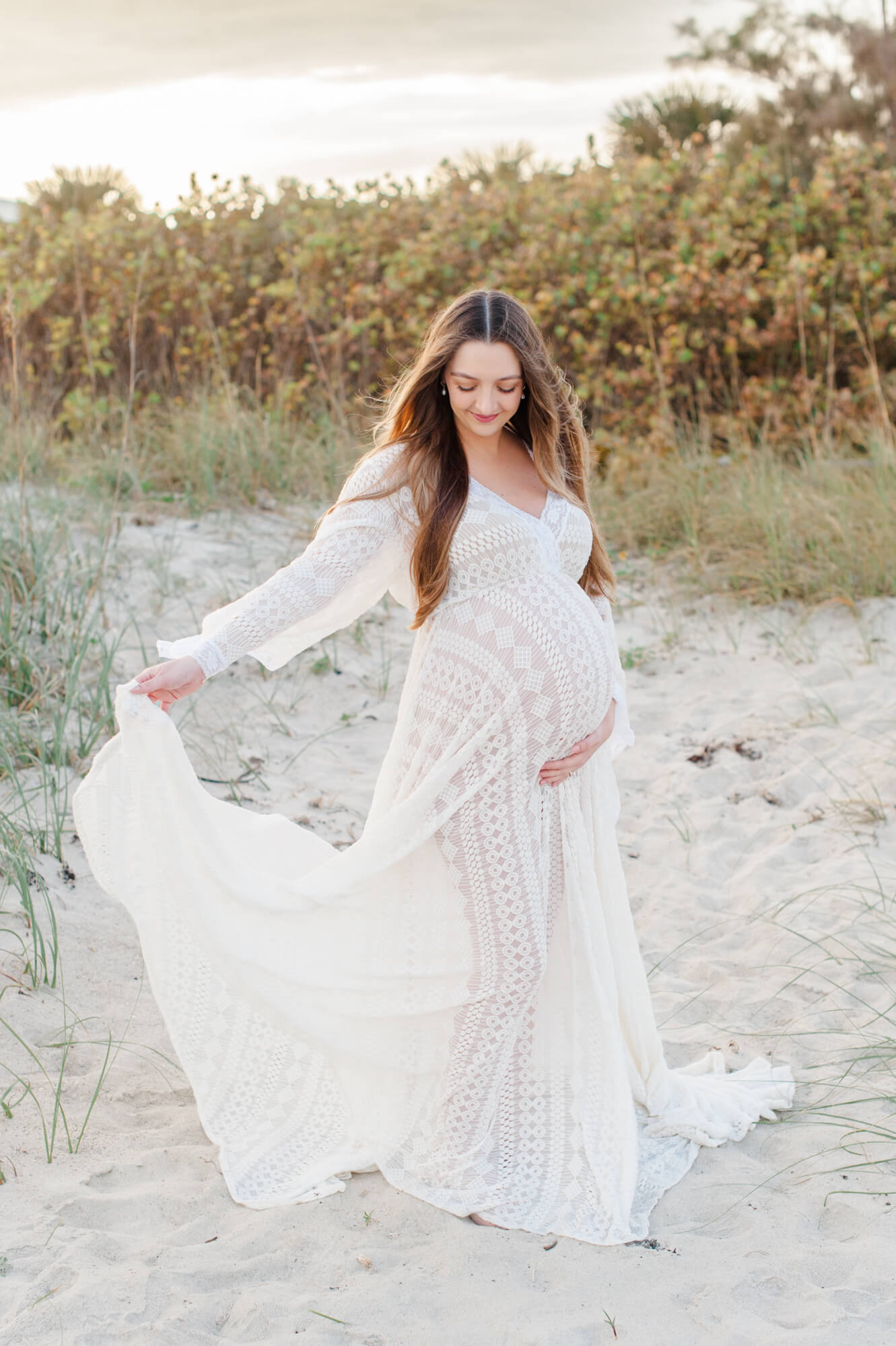 Pregnant mom holding her belly and waving her lace gown during golden hour near the dunes Tree of Life Birth Center