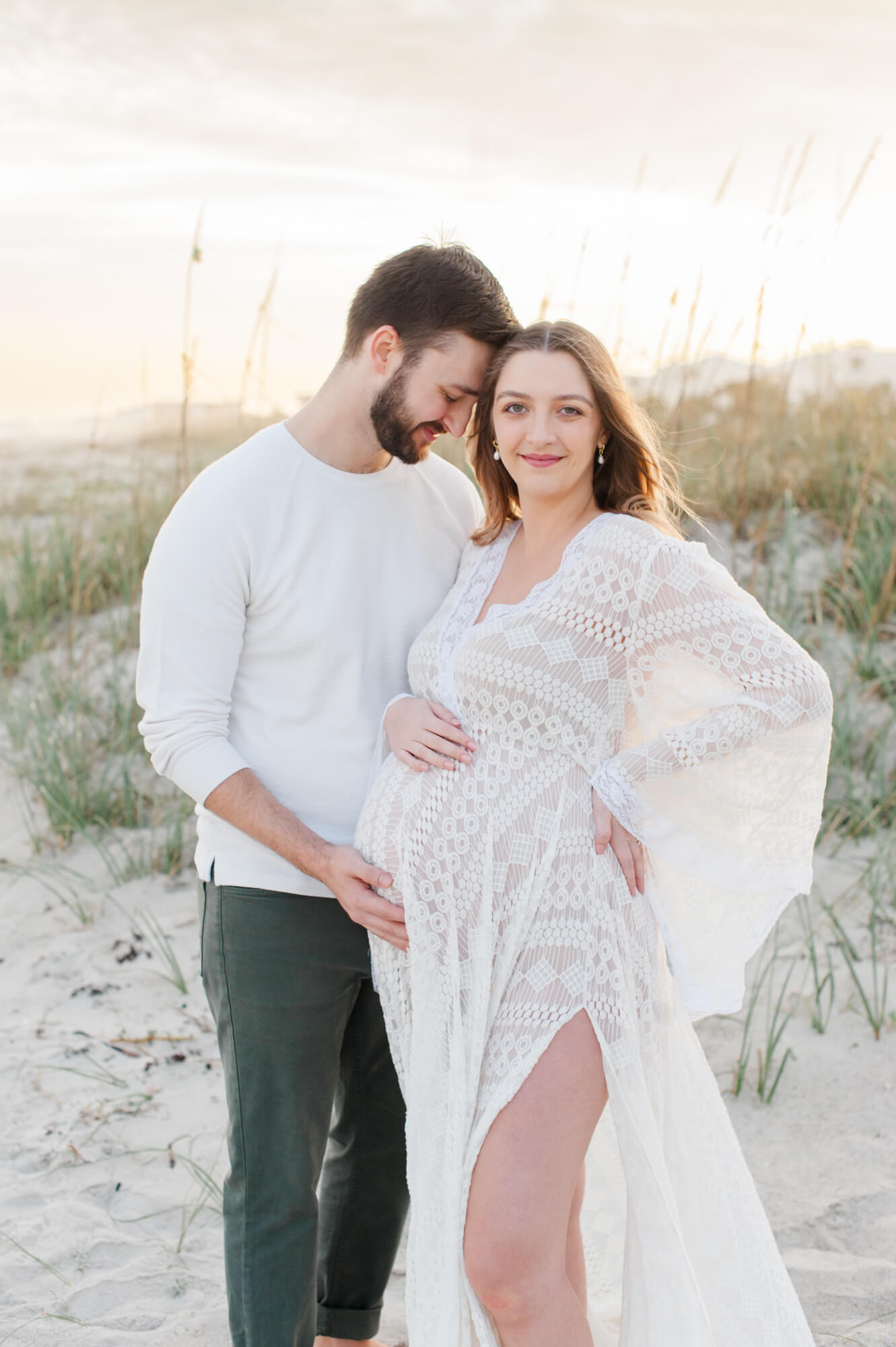 Expectant mother stares into the camera at sunset holding her belly with her husband. 