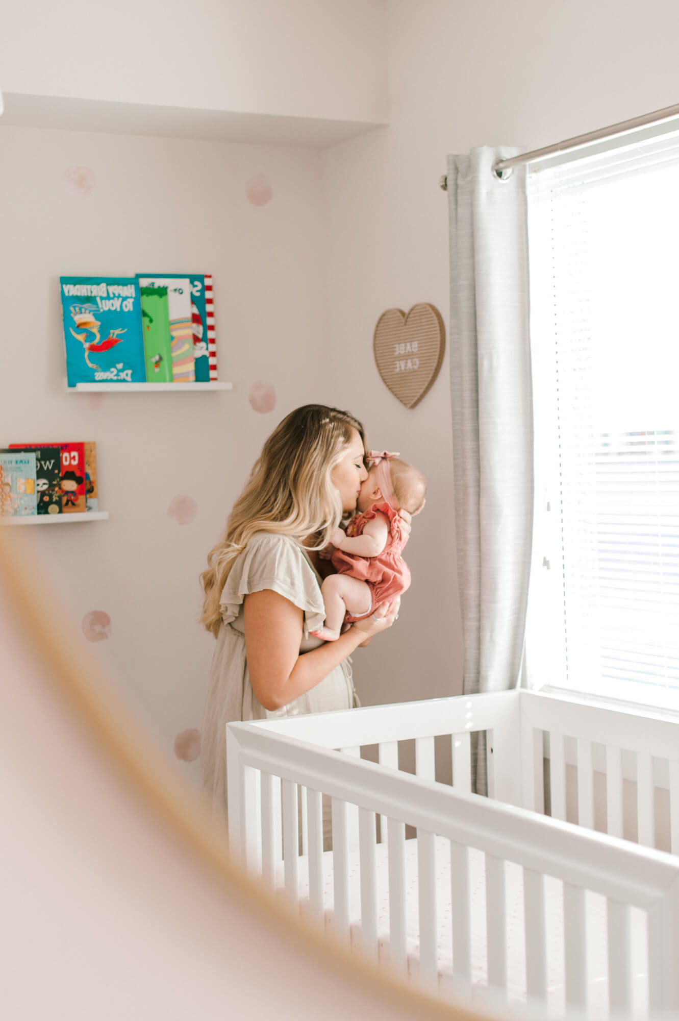 Mom holds daughter and kisses her nose next to the window in her bedroom.