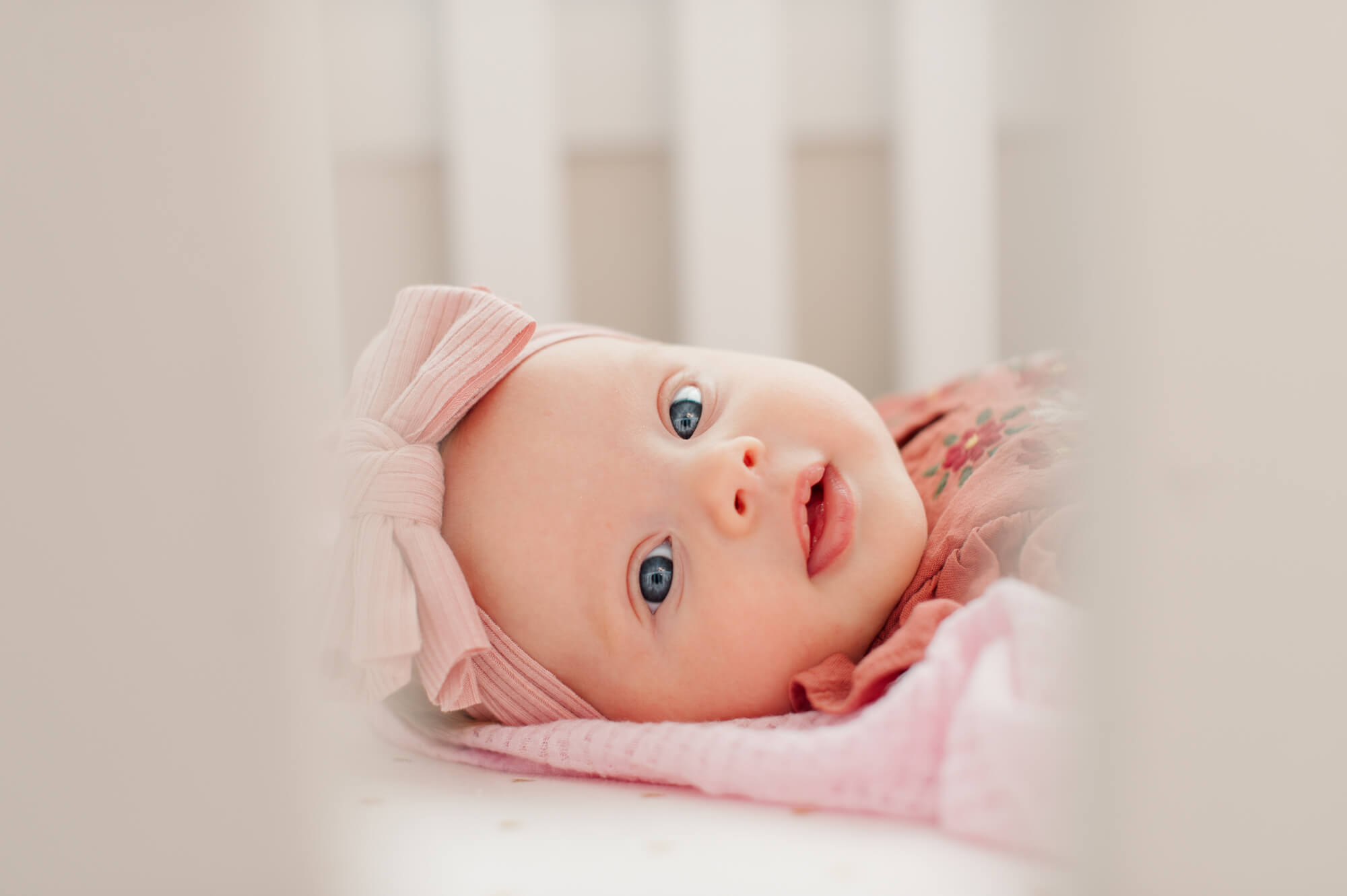 Newborn looks into the camera through the crib rails wearing all pink with bright blue eyes
