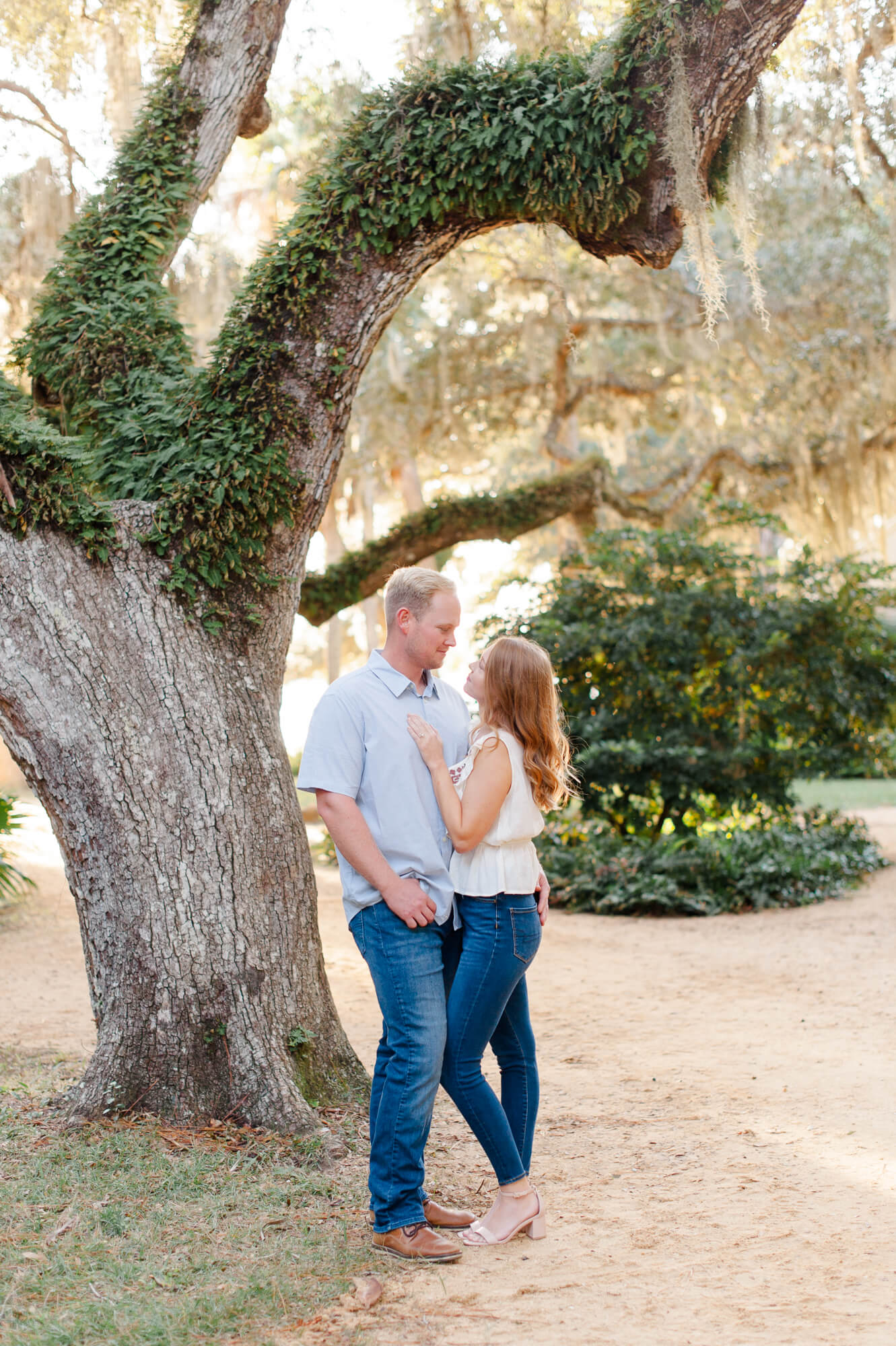 Couple stands near an old oak tree covered in greenery embracing each other with the sun setting behind them.
