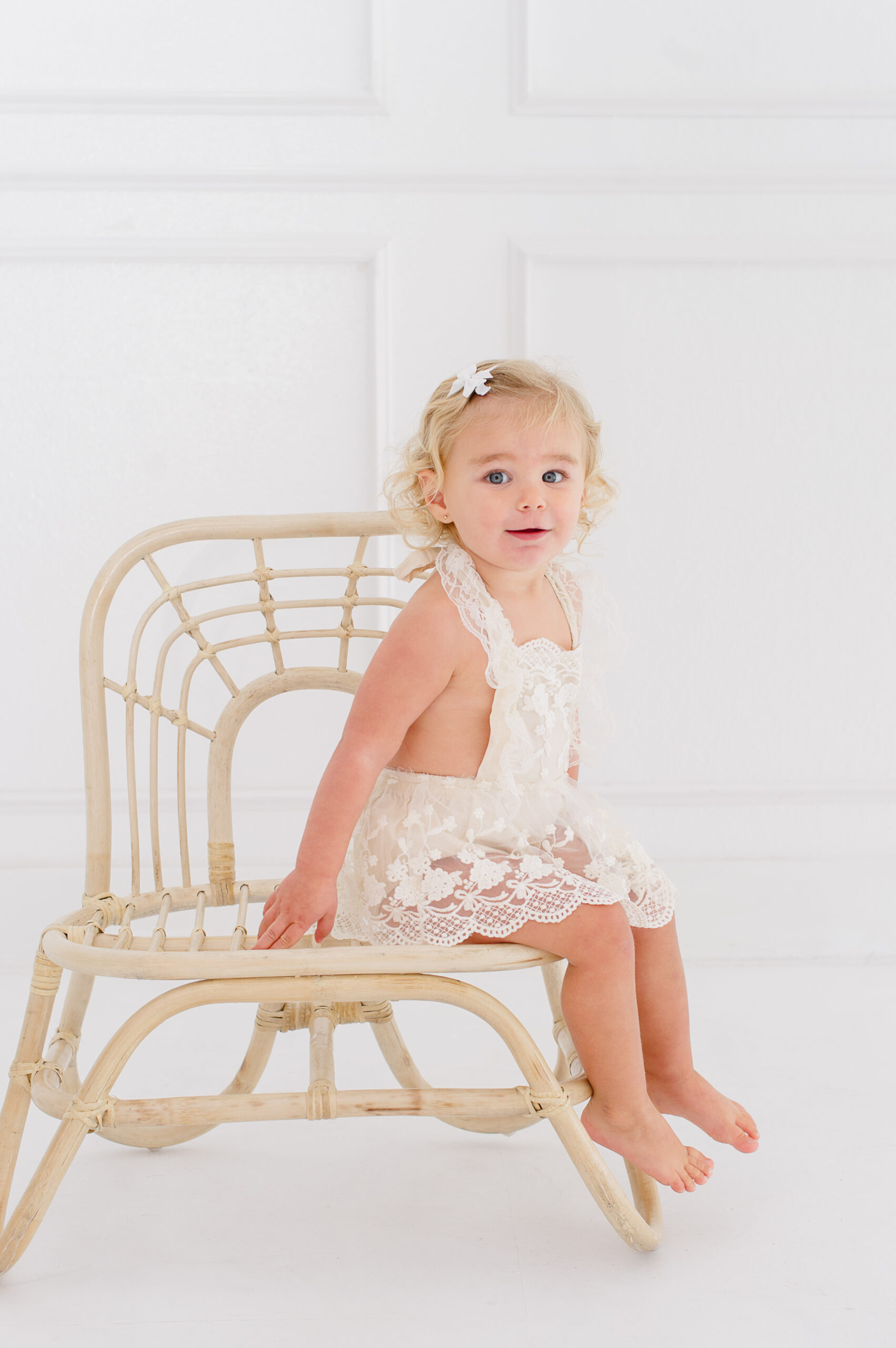 Sweet baby girl sitting in a wicker chair in a natural light studio in Orlando florida wearing a lace romper baby boutiques Orlando