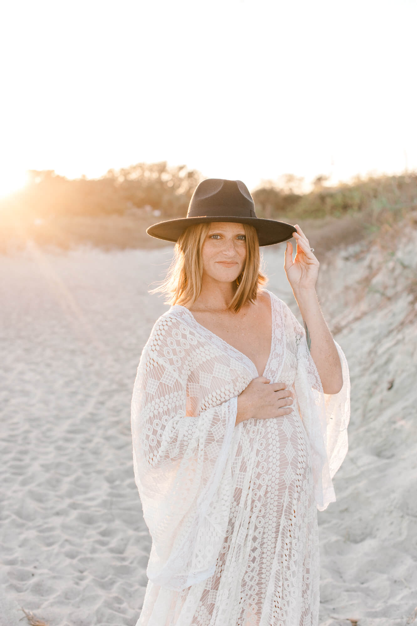 New mom places hand on her belly and smiles softly while holding her hat on the beach. 