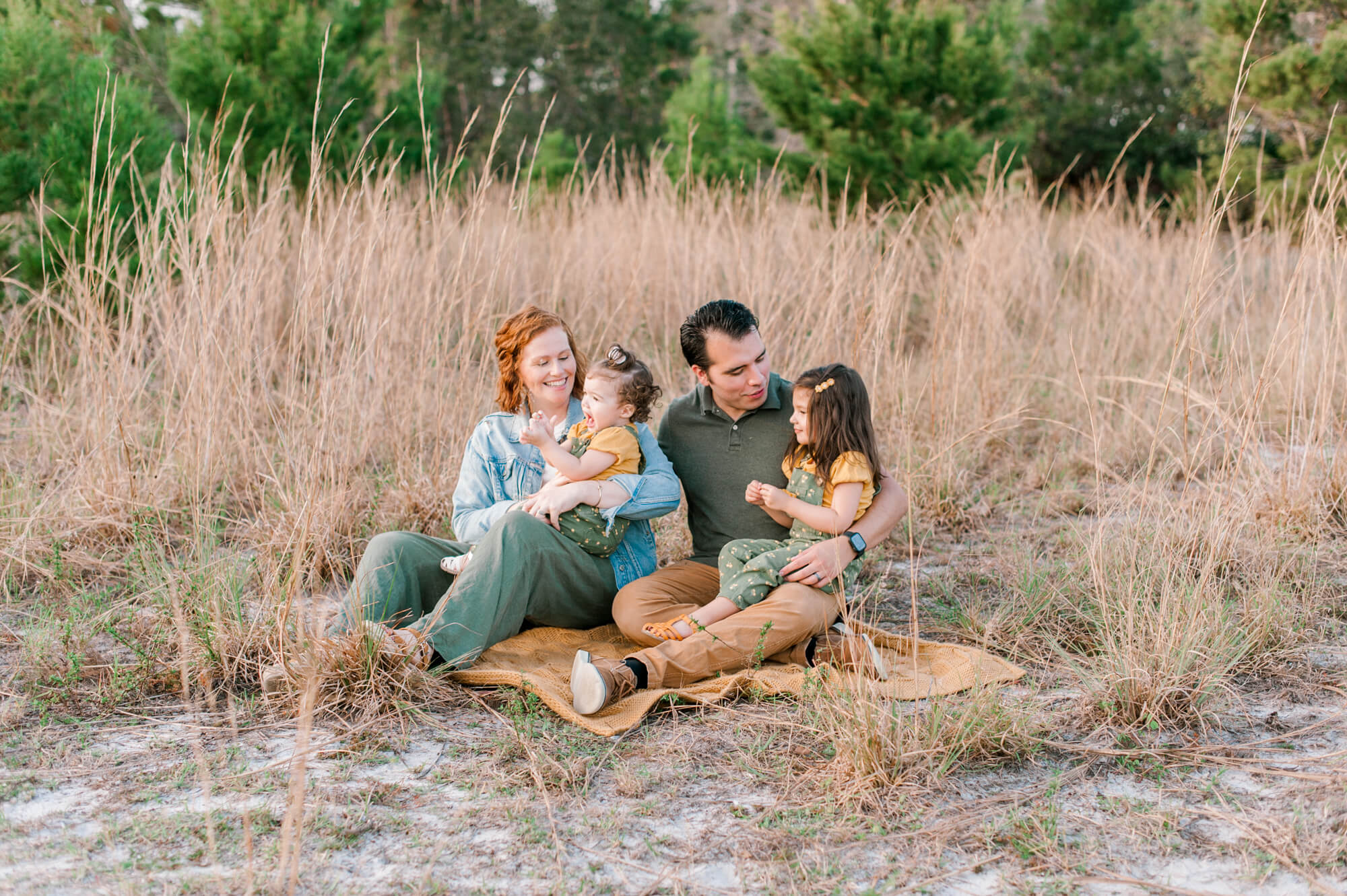 Family sitting on a blanket in tall grass tickling the girls. 