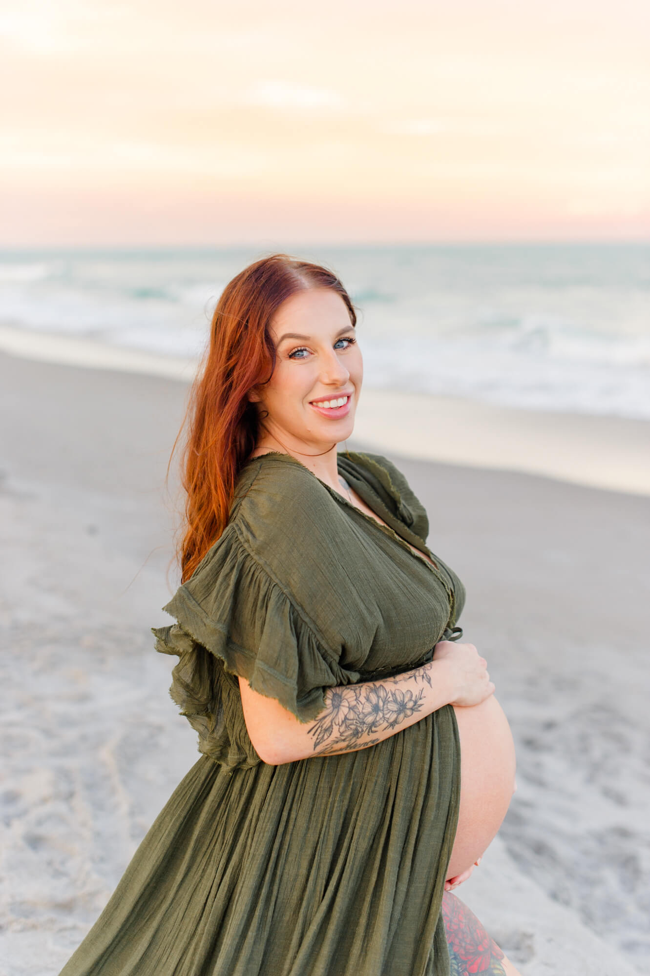 Beautiful expectant mother smiles at the camera while wearing a green maternity dress and standing near the shoreline.