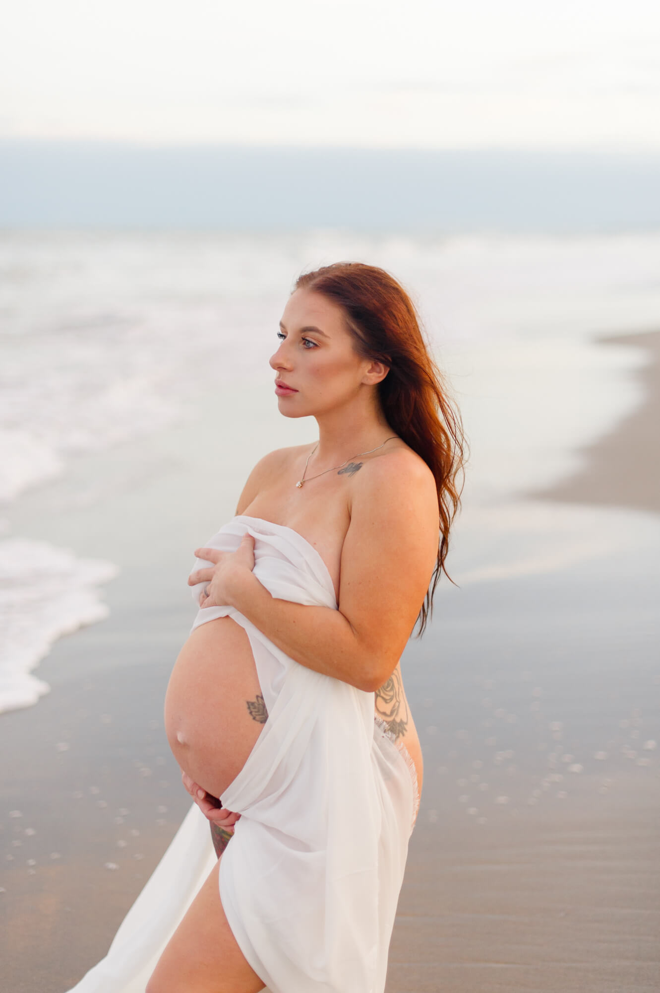 Beautiful new mother stands near the shoreline covered in white chiffon watching the waves crash while holding her belly at sunset 3D ultrasound Melbourne FL