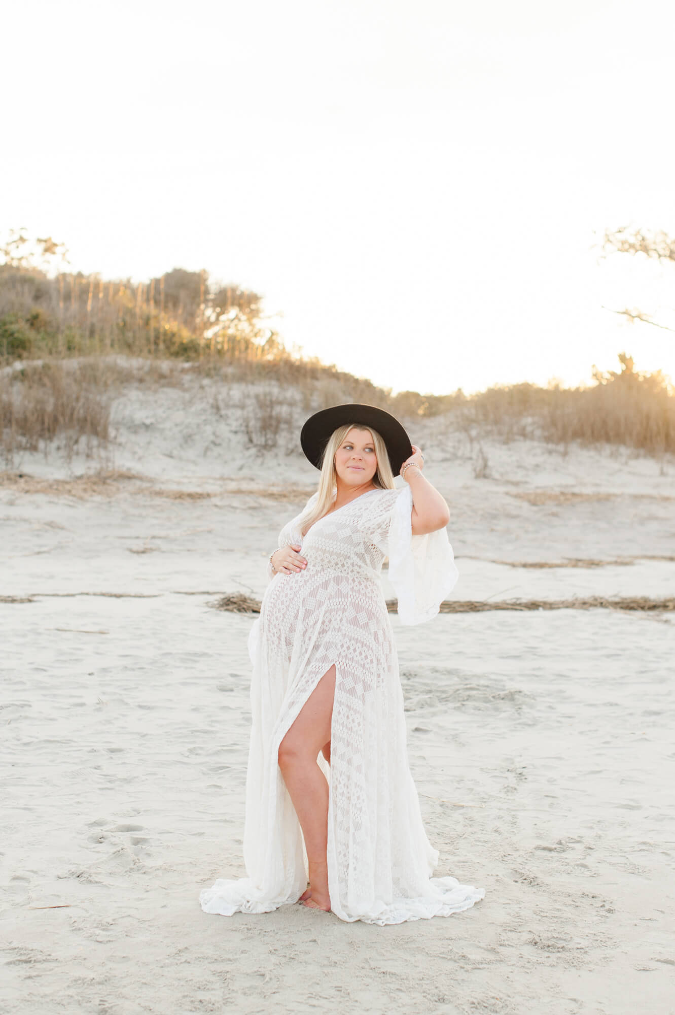 Beautiful new mother poses on the beach holding her wide brim hat and belly in a white lace gown!