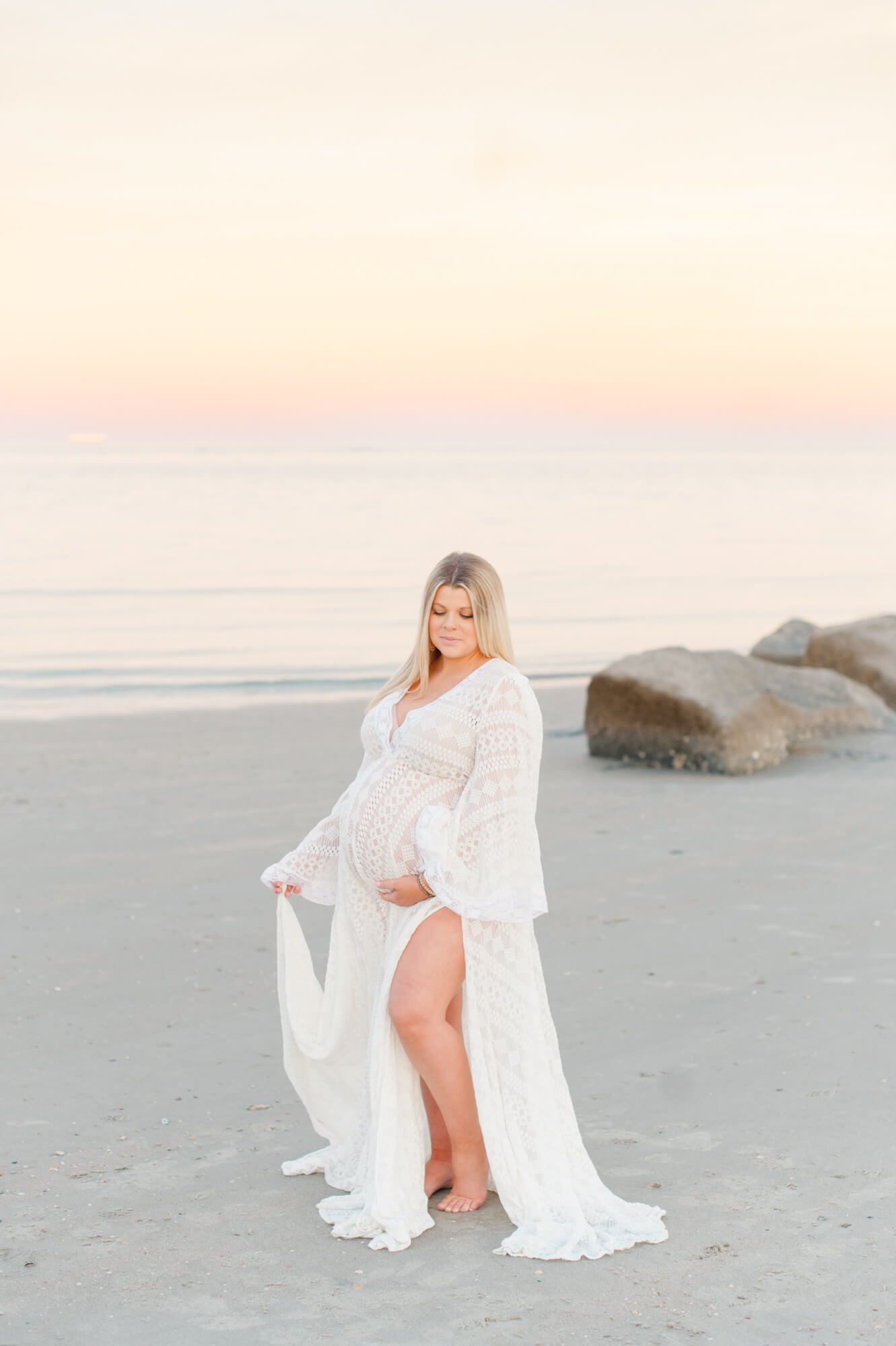 Stunning expectant mother stands holding her white lace gown and her belly during golden hour on the beach. Reflections OBGYN