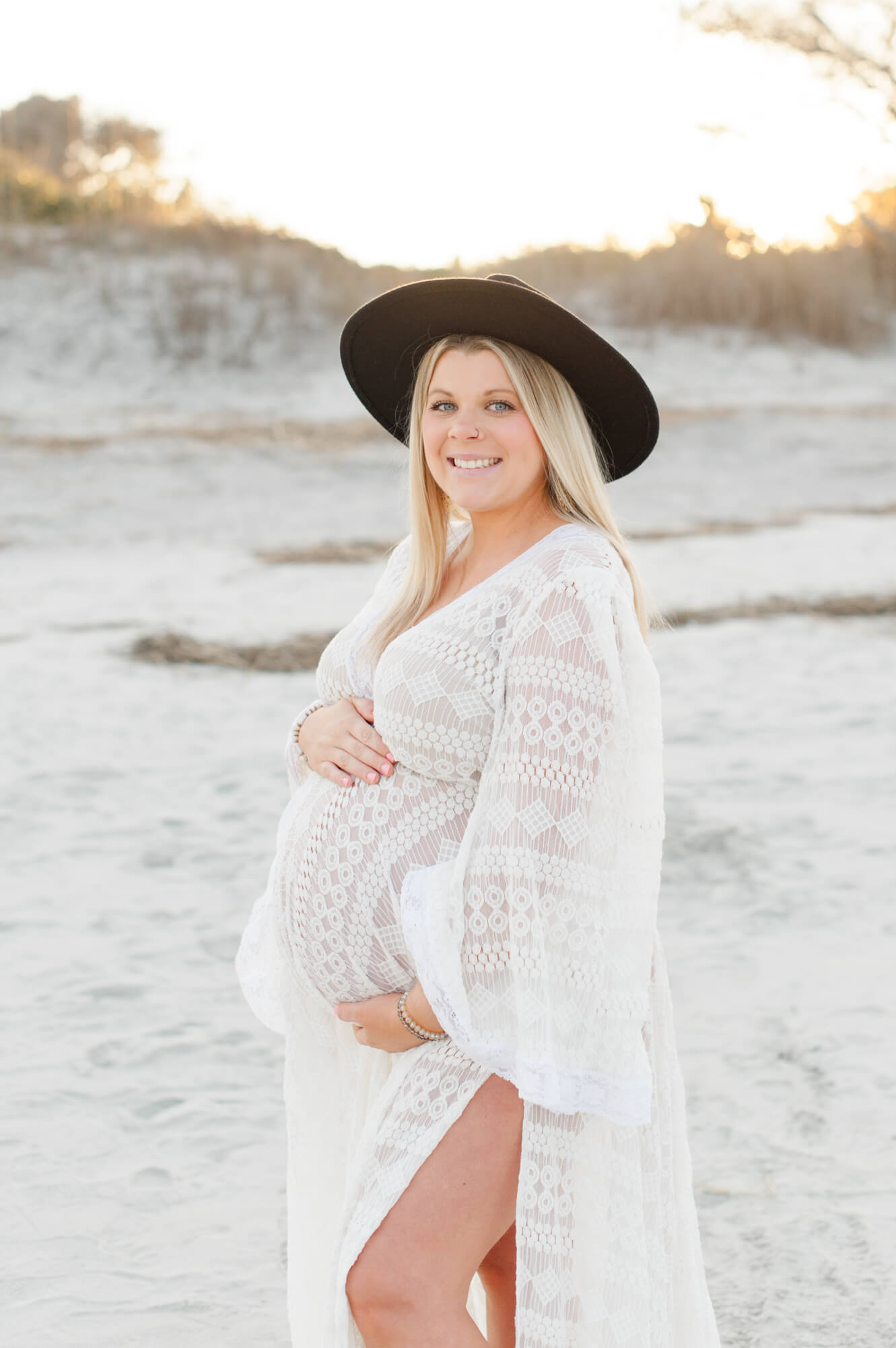 Expectant mother smiles at the camera during sunset while holding her belly in a white lace gown!