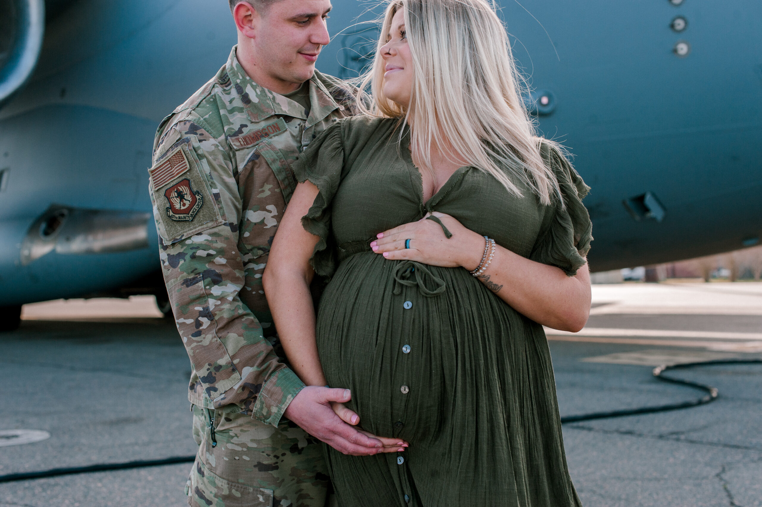 Close up of new mom and dad in front of military aircraft, mom holding her belly while thinking about Hibiscus Women's Center.