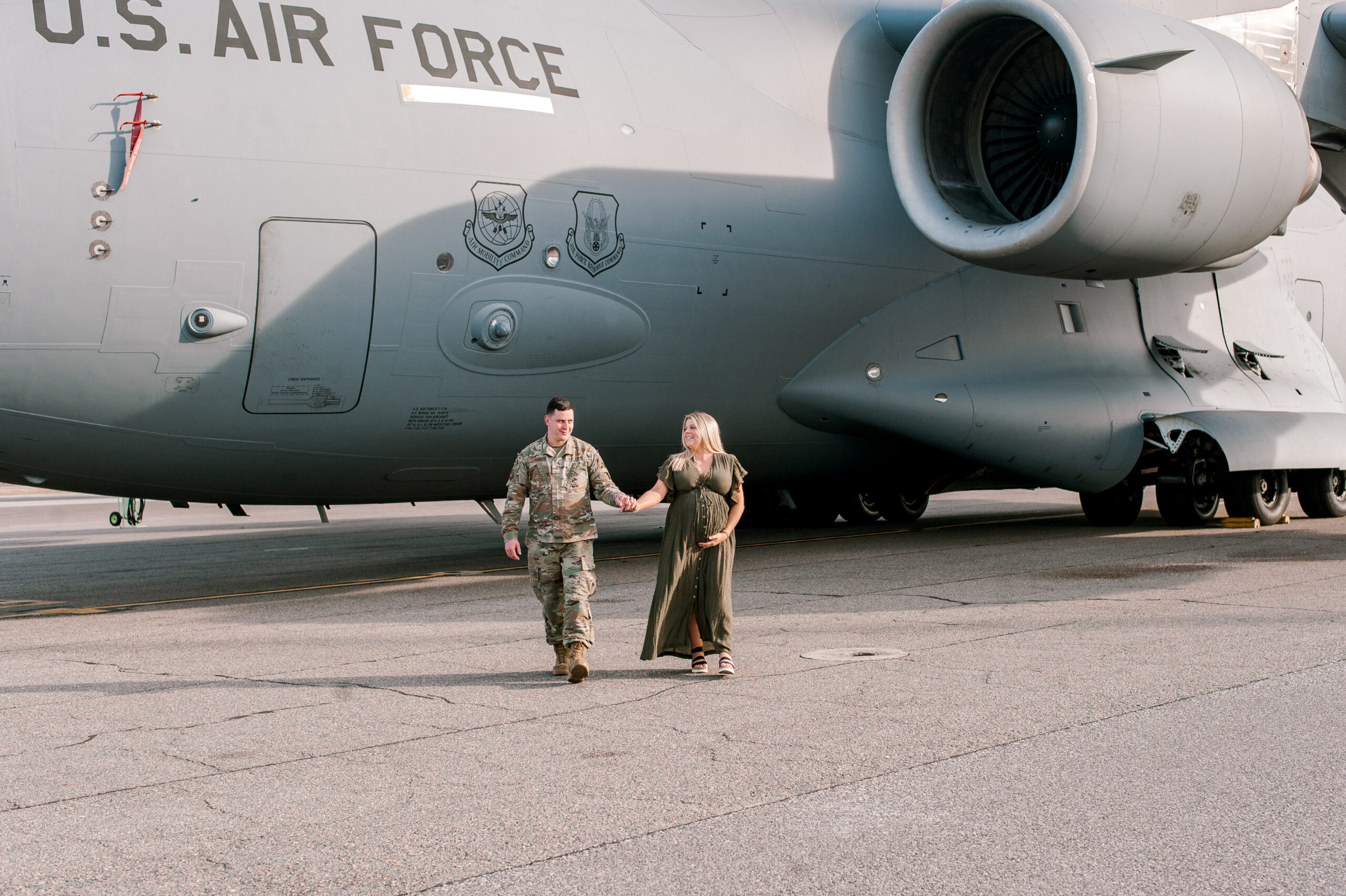 New parents walk hand in hand on the flight line in front of a military aircraft while mom holds her belly and thinks about how incredible the hibiscus women's center is. 