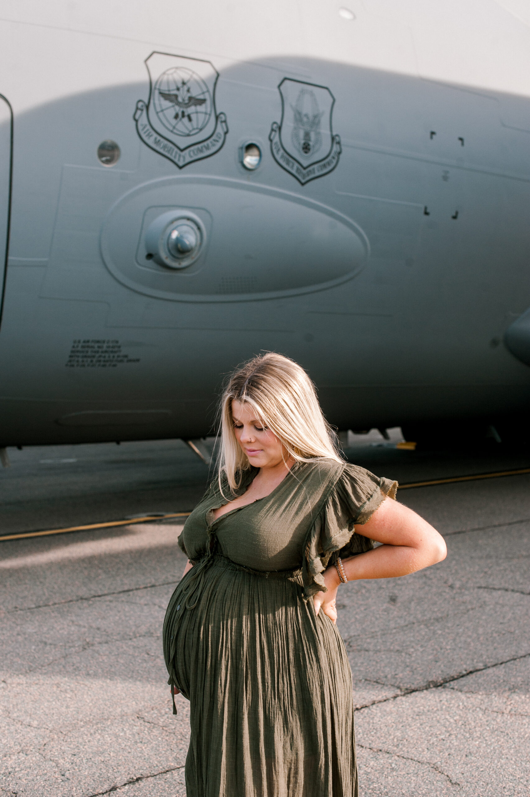 Soon to be new mama holding her belly and looking down at her baby in front of a military aircraft on the flight line. 