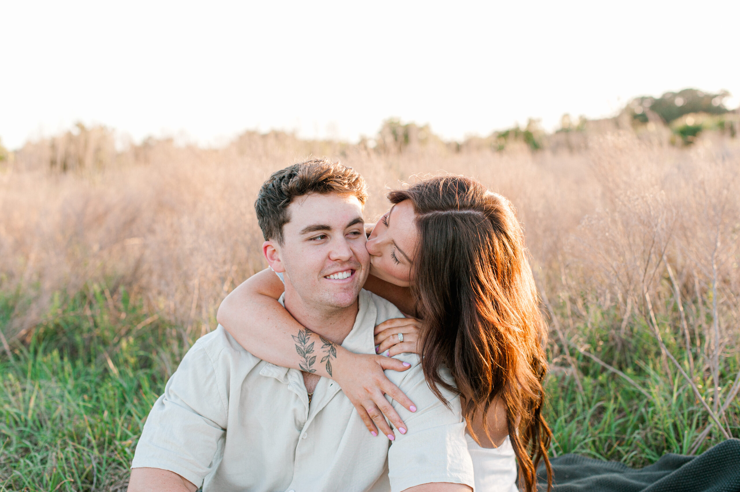 Girl hugging her boyfriends neck while kissing his cheek and whispering in his ear at sunset in tall grass. 