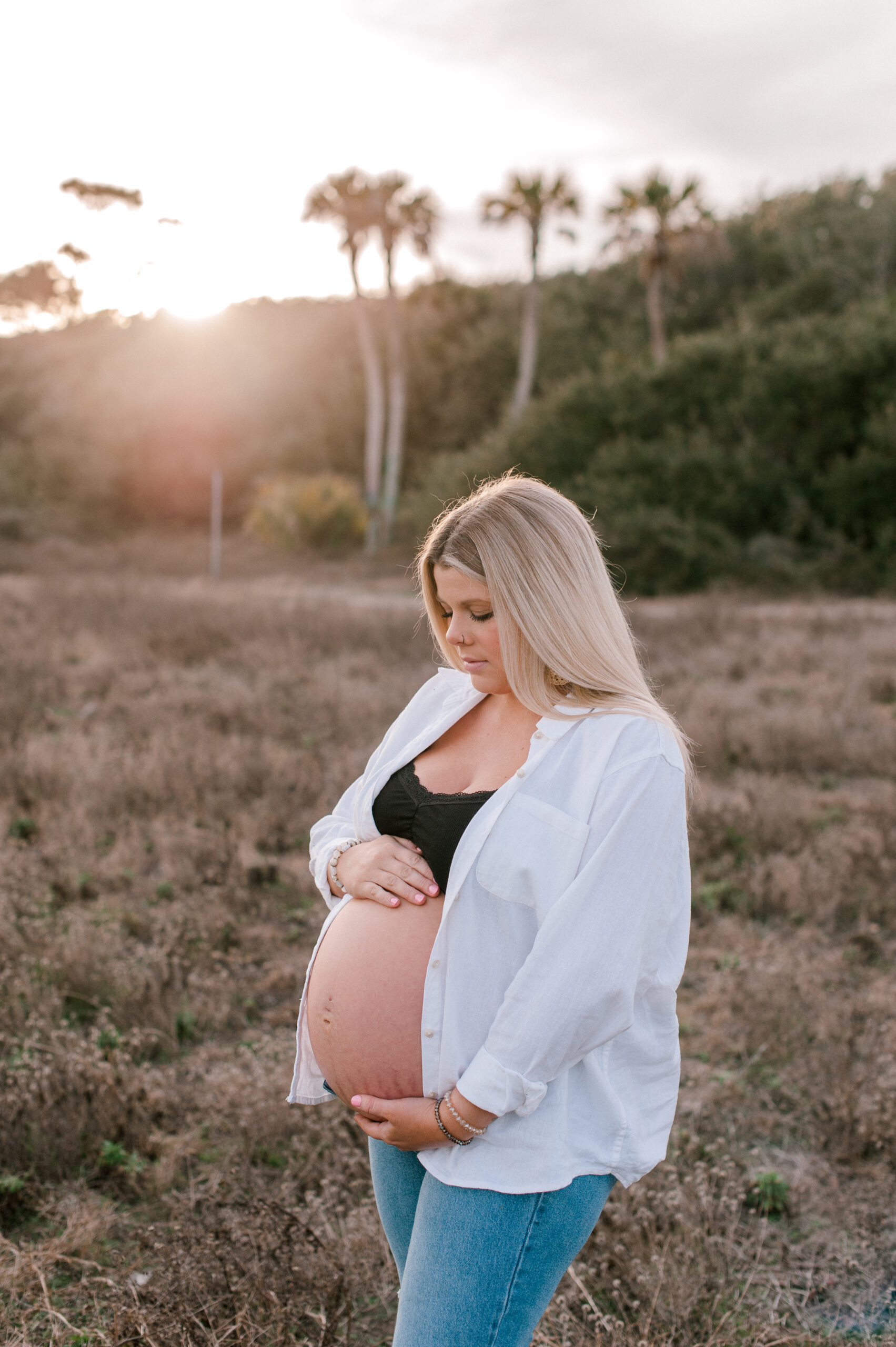 New mom stands in a field at sunset holding her belly with both hands and glancing down at her baby. 