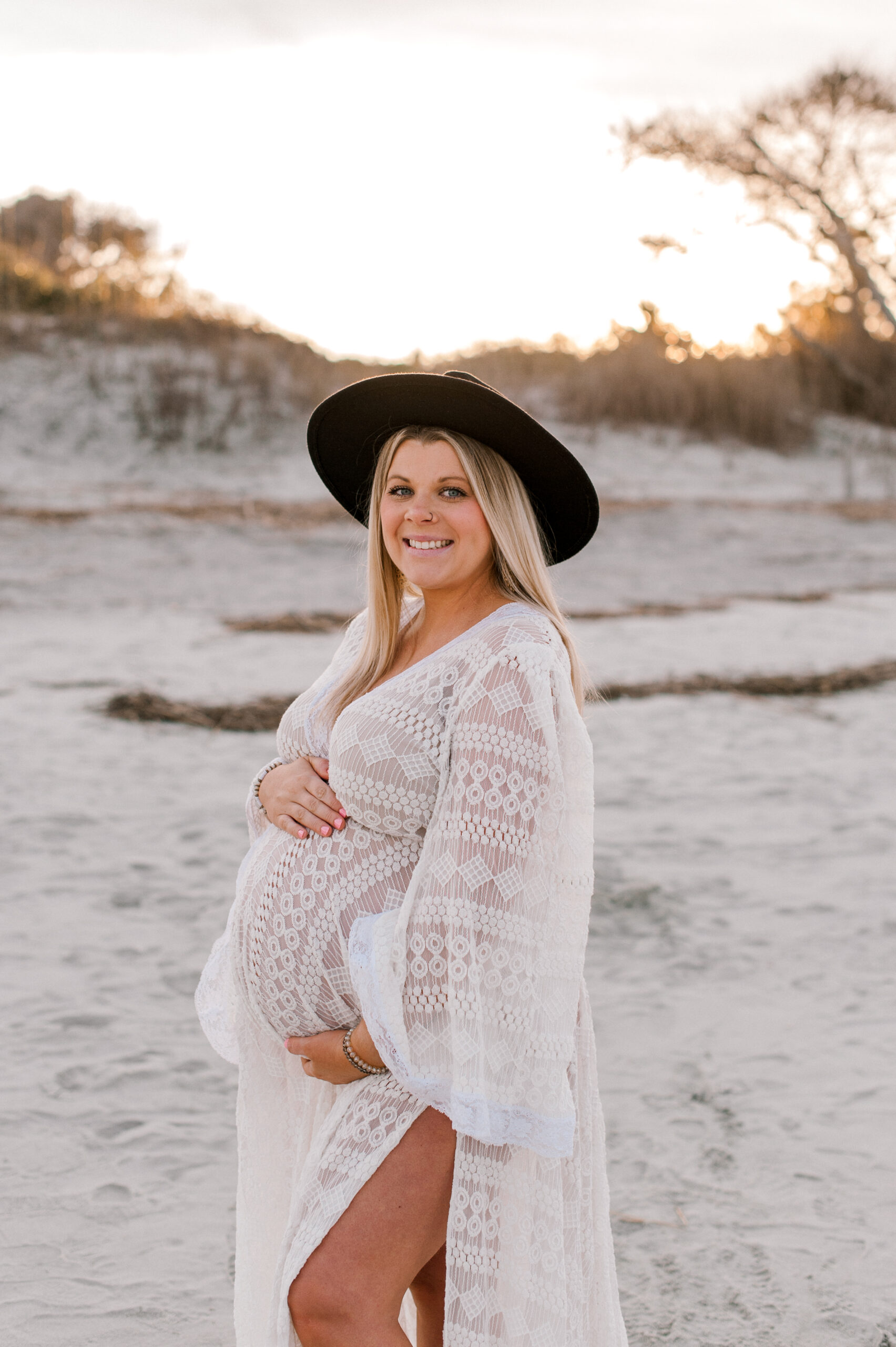 New mama is glowing at sunset, smiling at the camera while holding her belly with both hands. 