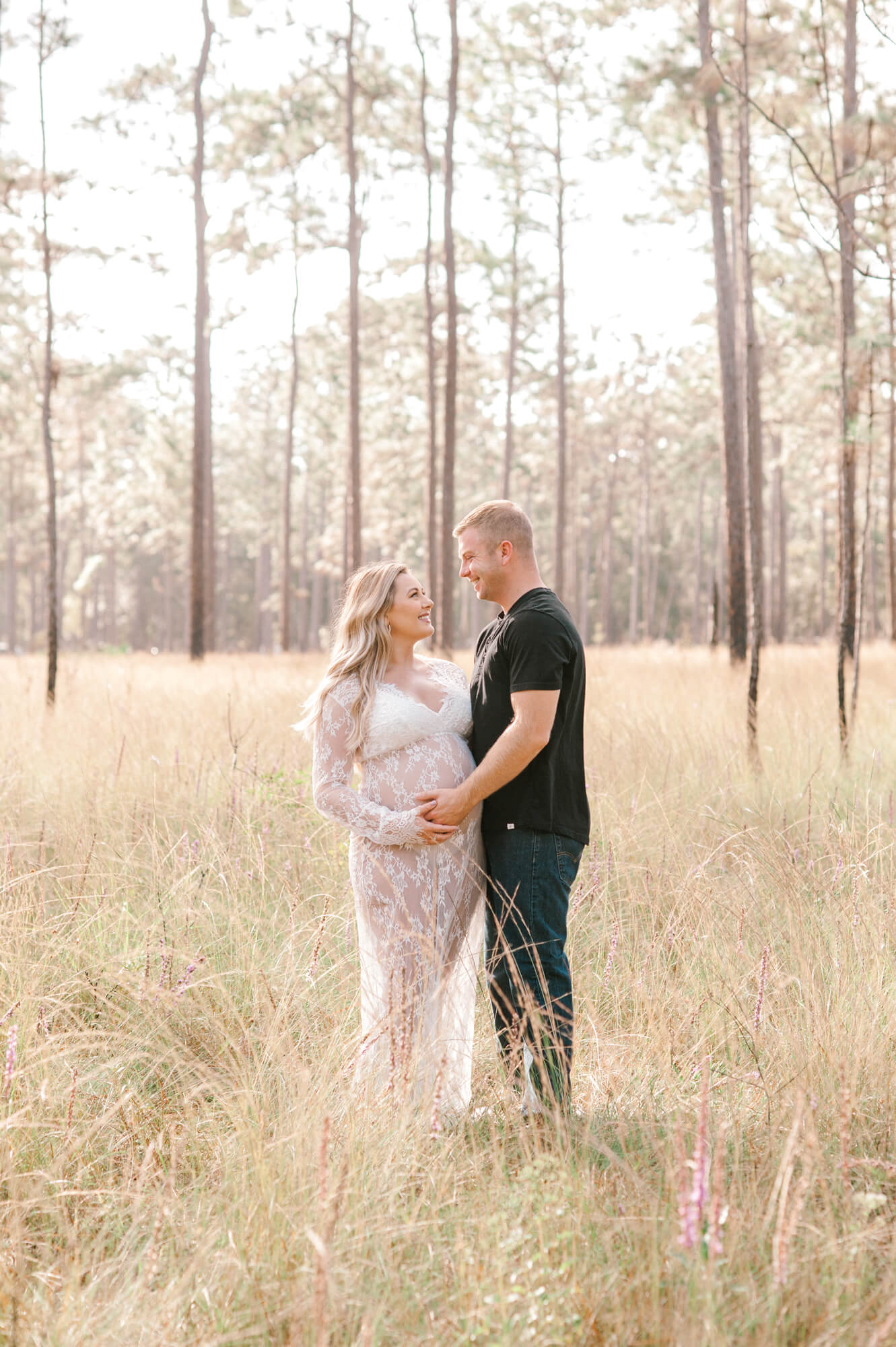 Couple stands in a field of tall grass surrounded by trees holding the moms belly thinking about their new daughter.