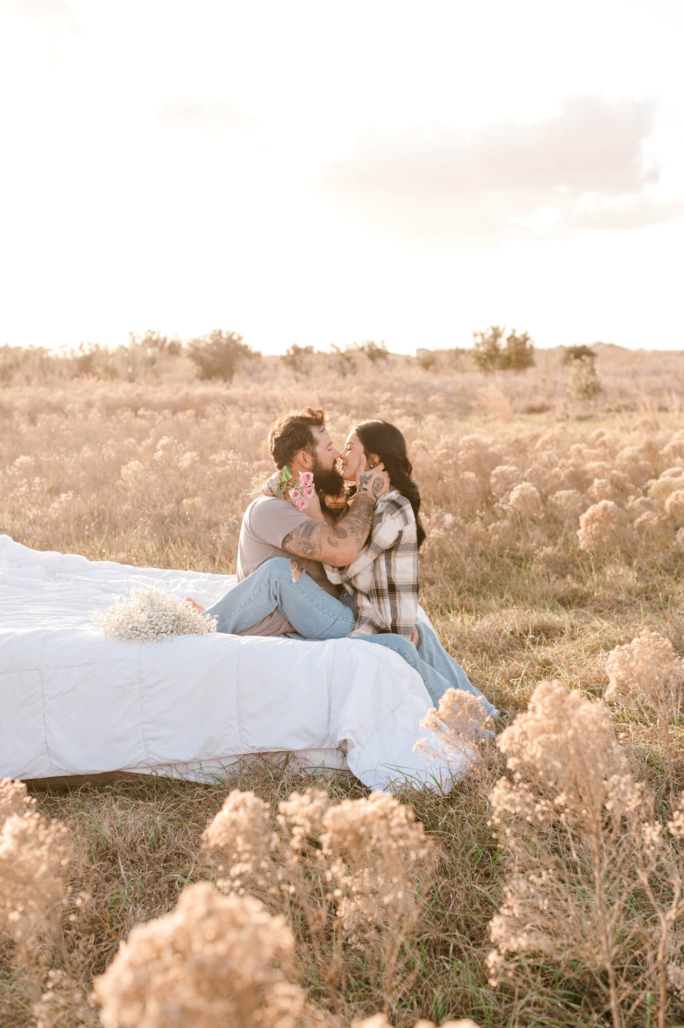 Wife sitting in her husbands lap facing him while going in for an intimate kiss at sunset in the middle of a beautiful field. 