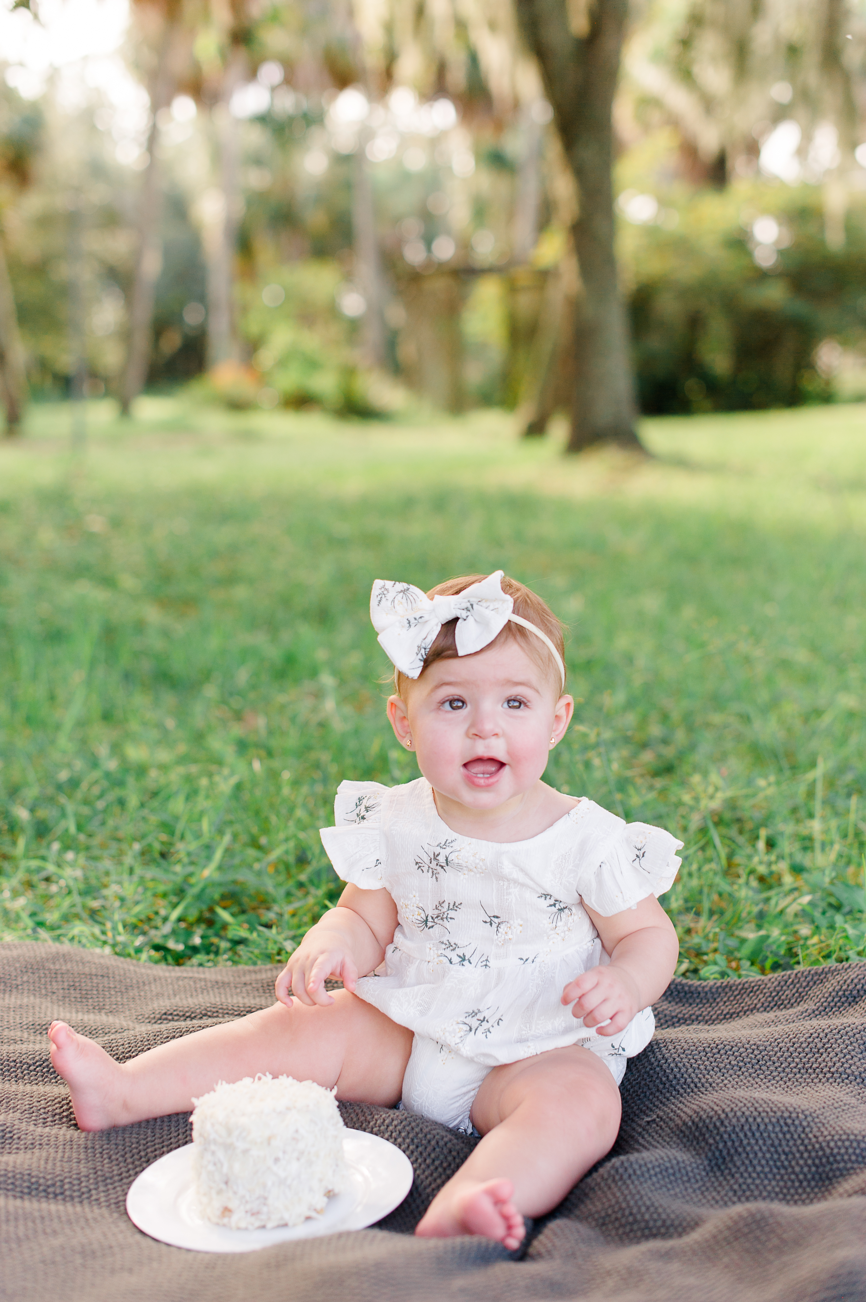 Twinkle Toes Nanny Agency would be perfect for this babygirl sitting on a comforter wearing a lace romper and bow while smiling at the camera.