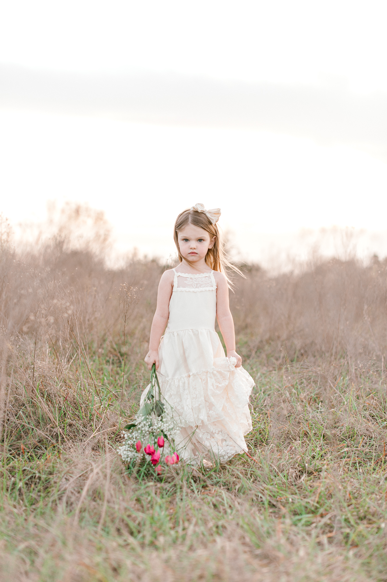 Toddler girl walking through a tall grass field holding a bouquet of flowers. 