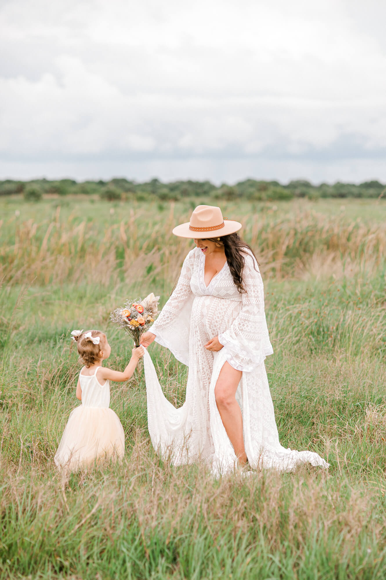 Toddler girl hands mom a bouquet wildflowers in a field during the moms maternity session.