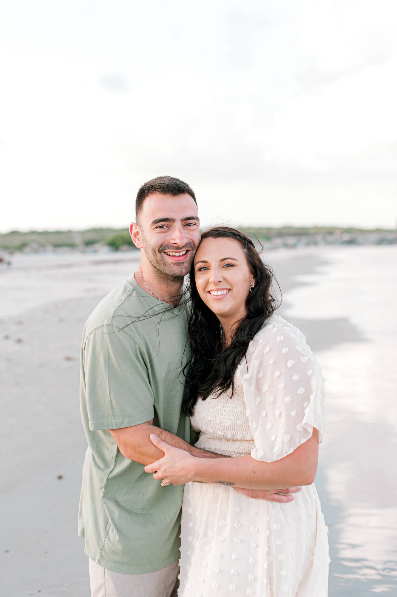Photo of the newlyweds smiling and cuddling close on the beach at sunset during their extended family beach session.