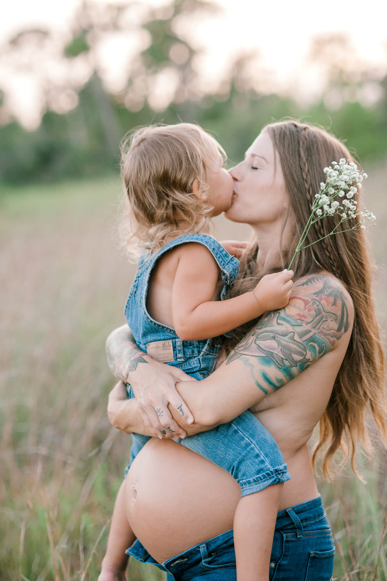 Pregnant mom and young daughter kissing during golden hour while standing in a tall grass field baby fairy tale