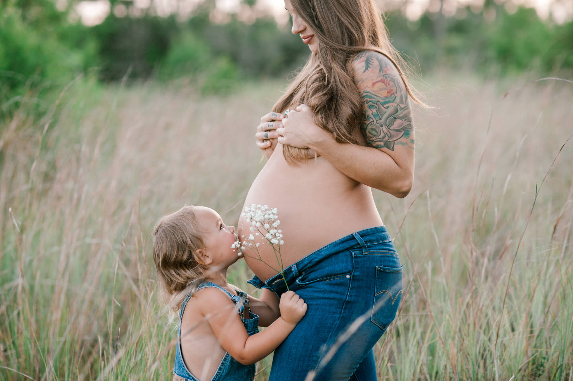 Expectant mom standing in tall grass covering her breasts while her young daughter kisses her pregnant belly at sunset baby fairy tale