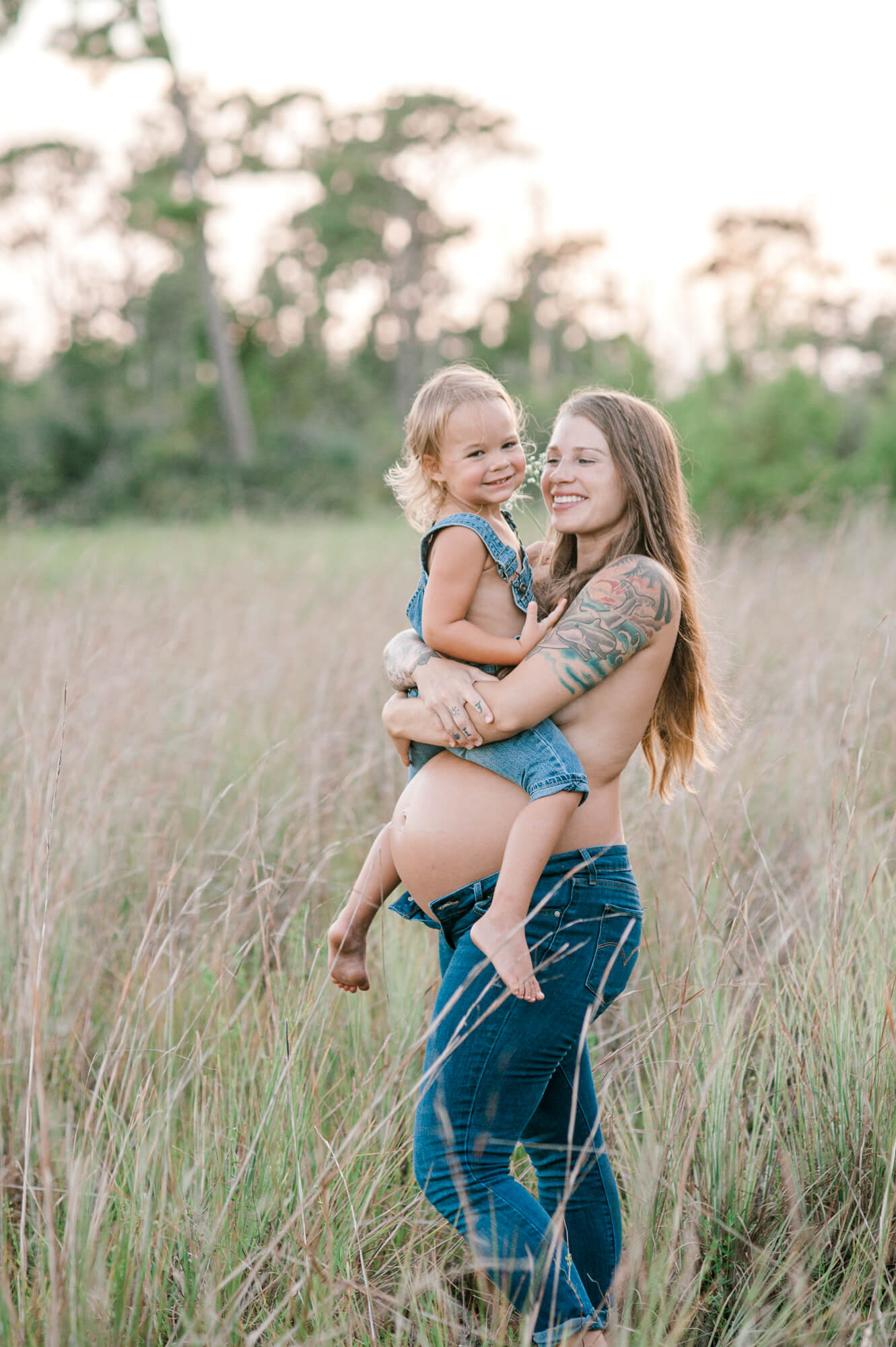 Mom holding daughter on her pregnant belly and laughing while standing in tall grass