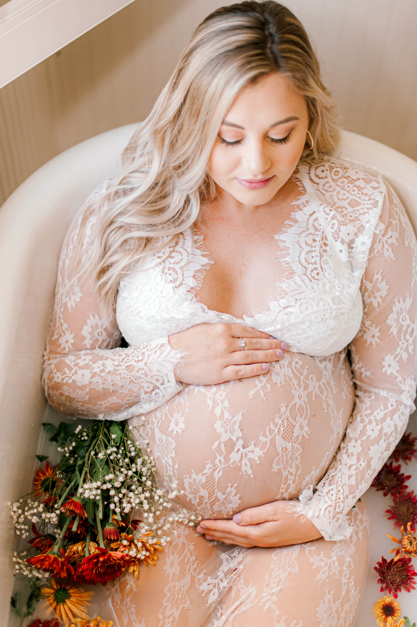Pregnant mama in lace dress sits in a milk bath holding her belly and looking down toward her babygirl 