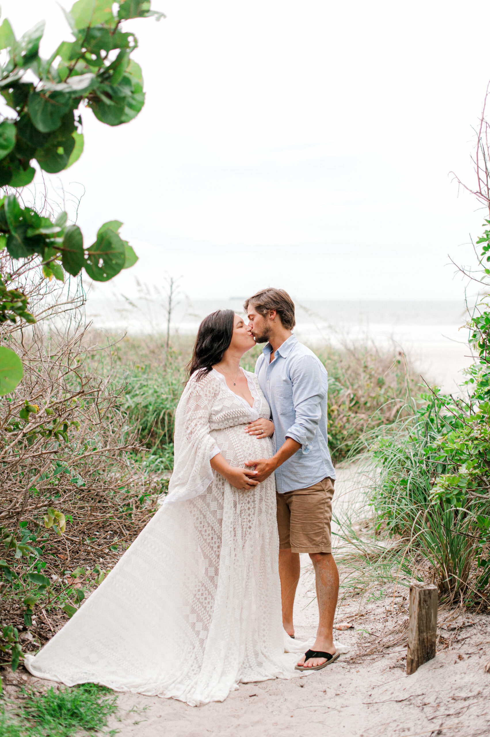 Soon to be new mom and dad kissing near the dunes with the beach in the background while holding her belly serendipity birthing