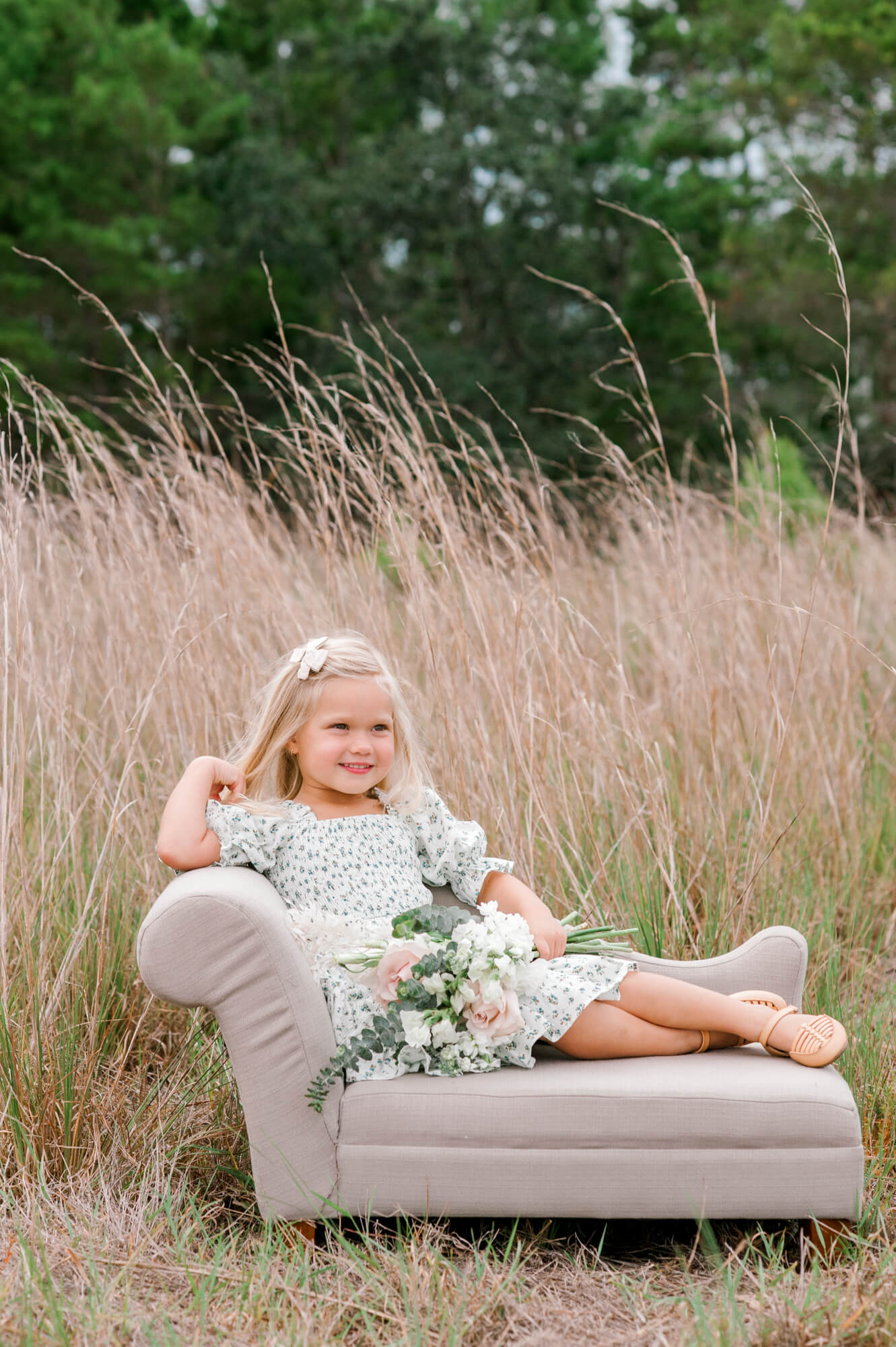 Young girl sitting on a miniature couch in a tall grass field at sunset holding a bouquet viera children's academy