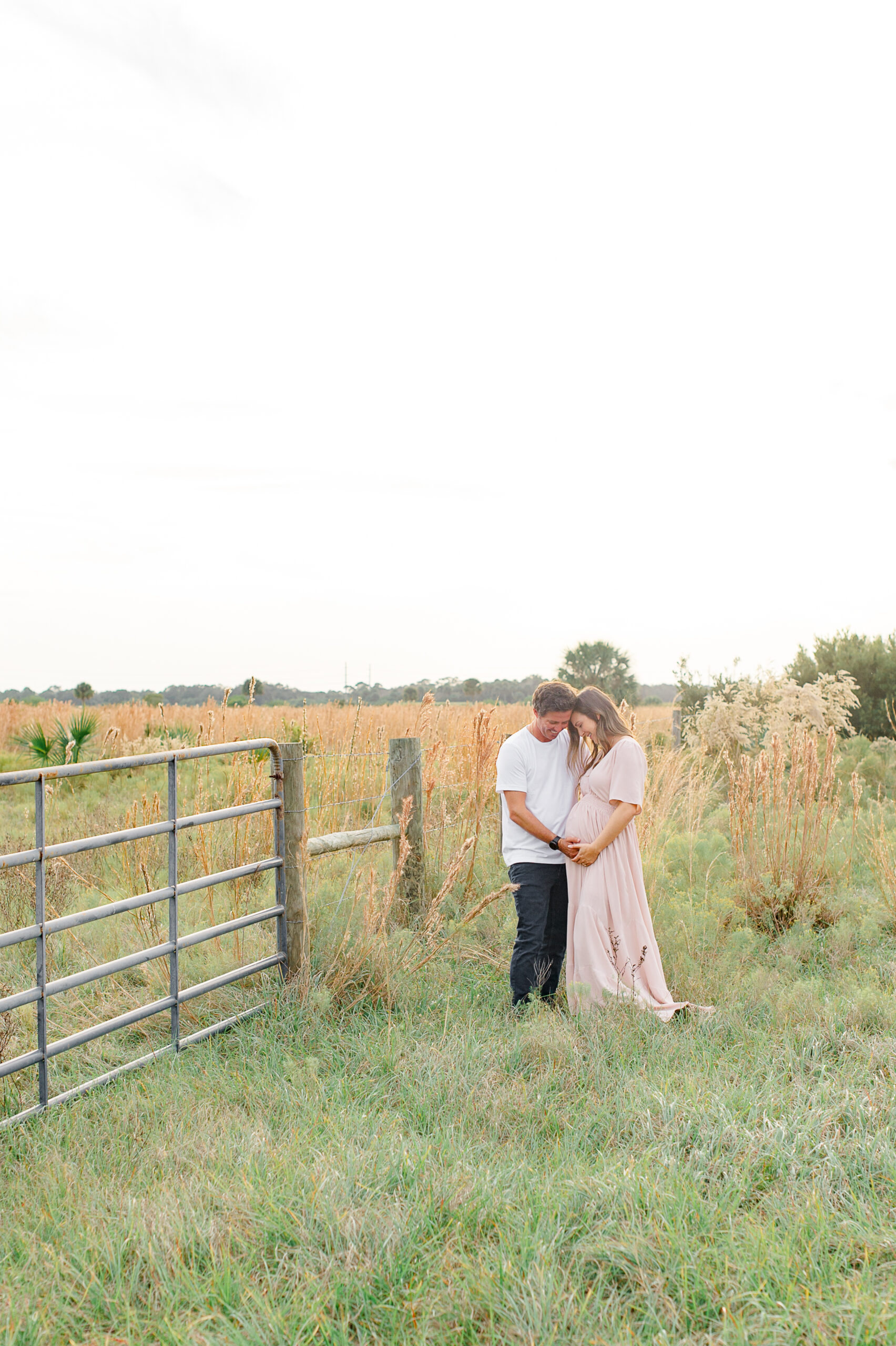 Parents stand in a tall grass field holding each other while looking toward moms pregnant belly during maternity photoshoot