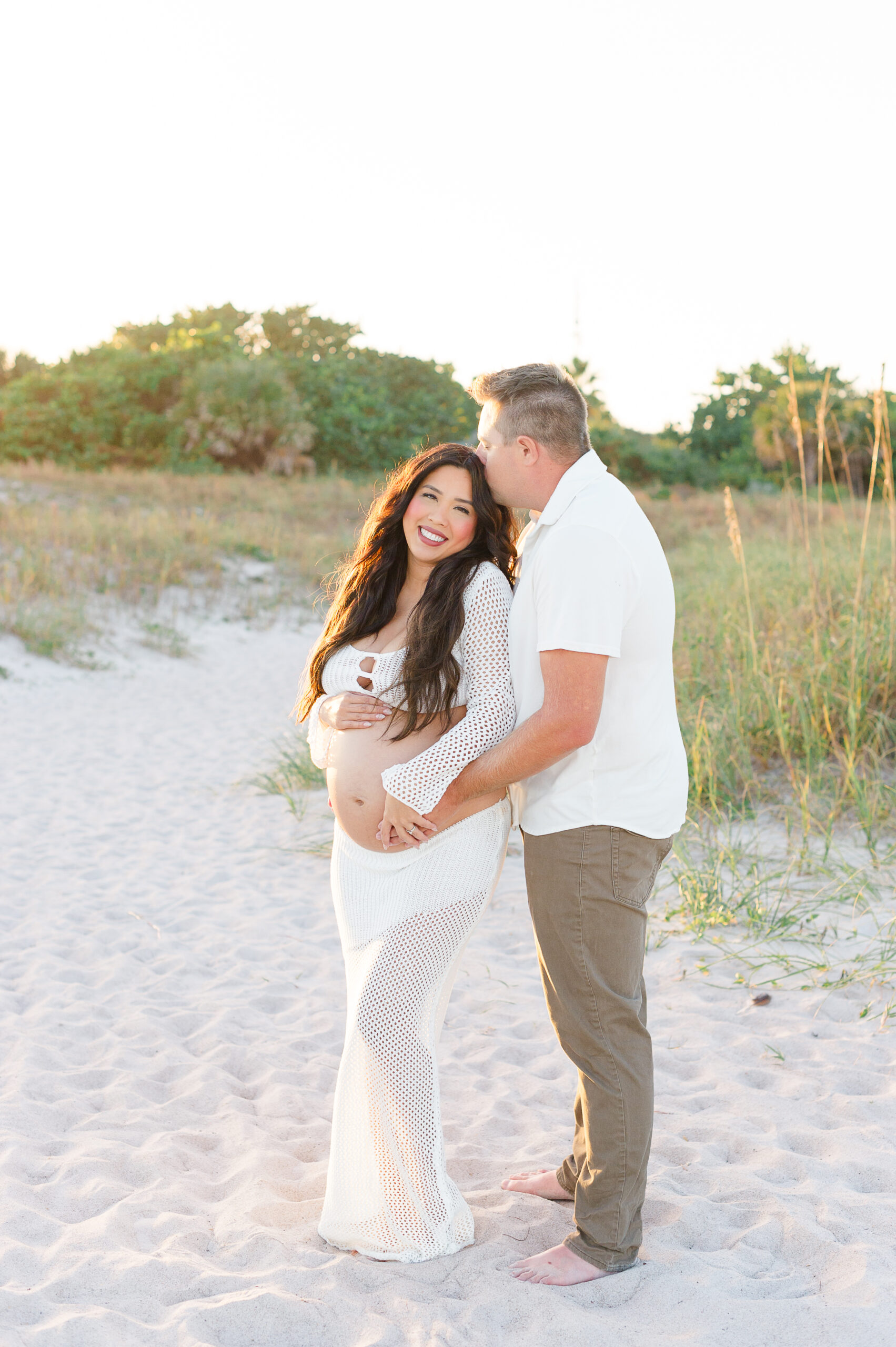 Expectant parents stand on the beach at sunset holding each other while dad kisses moms temple and they hold her pregnant belly.
