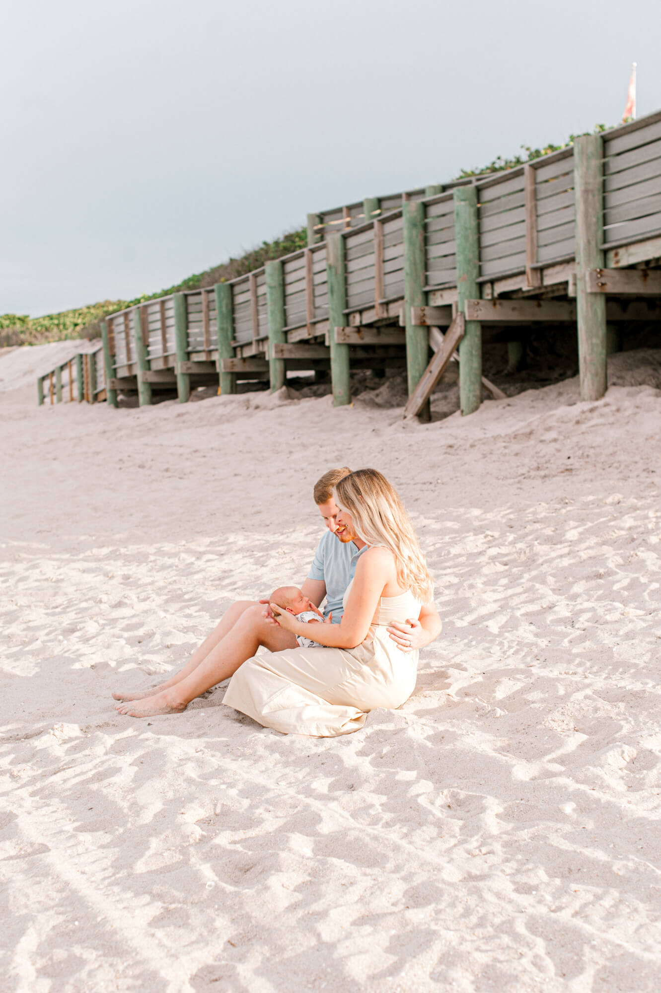 New parents sit in the sand cuddling their new baby boy close in front of a long wooden boardwalk at sunset during their newborn photoshoot Orlando baby stores