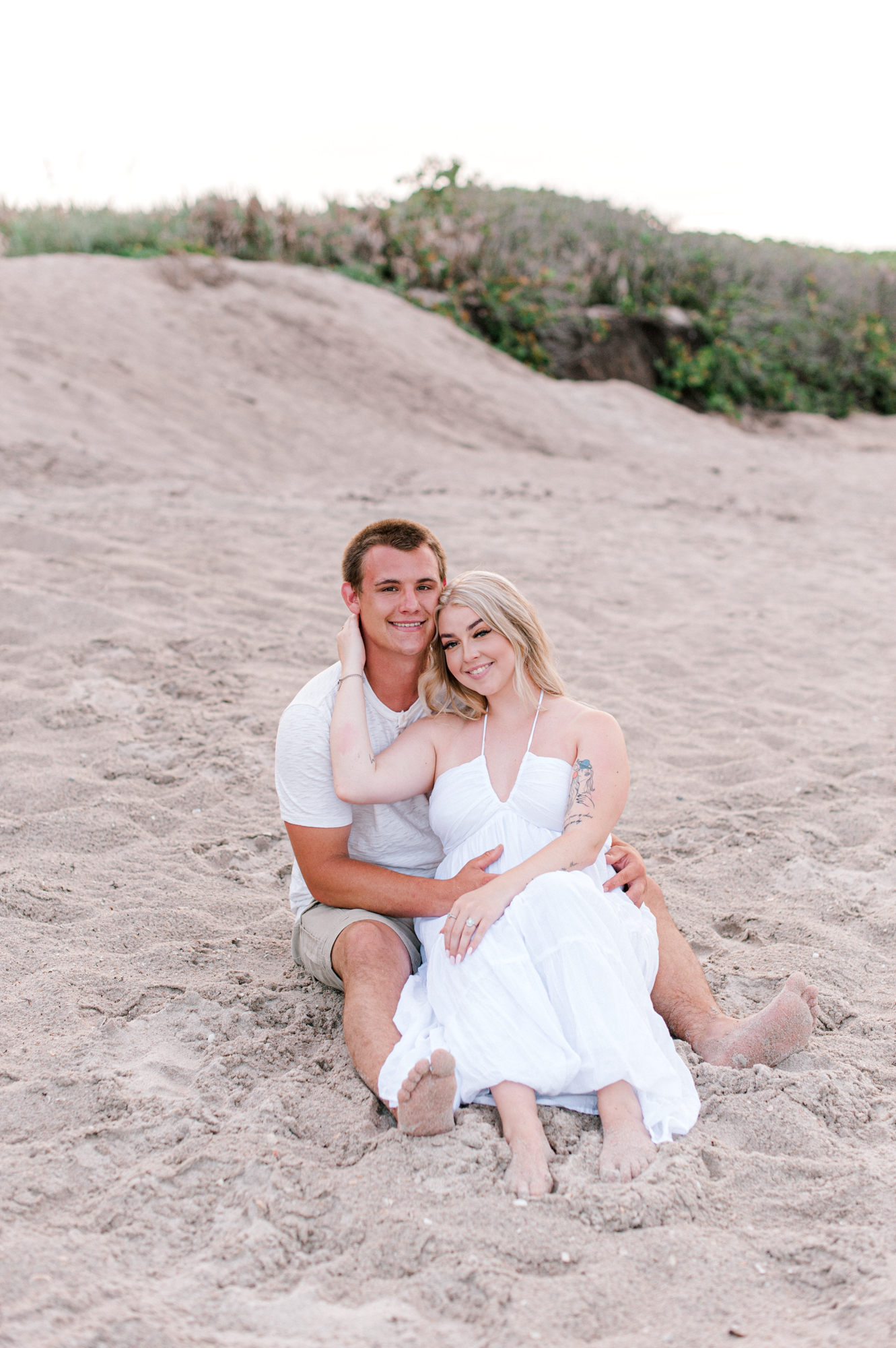 Couple sitting near the dunes during their engagement session on the beach smiling at the camera while cuddled close Orlando hair salons