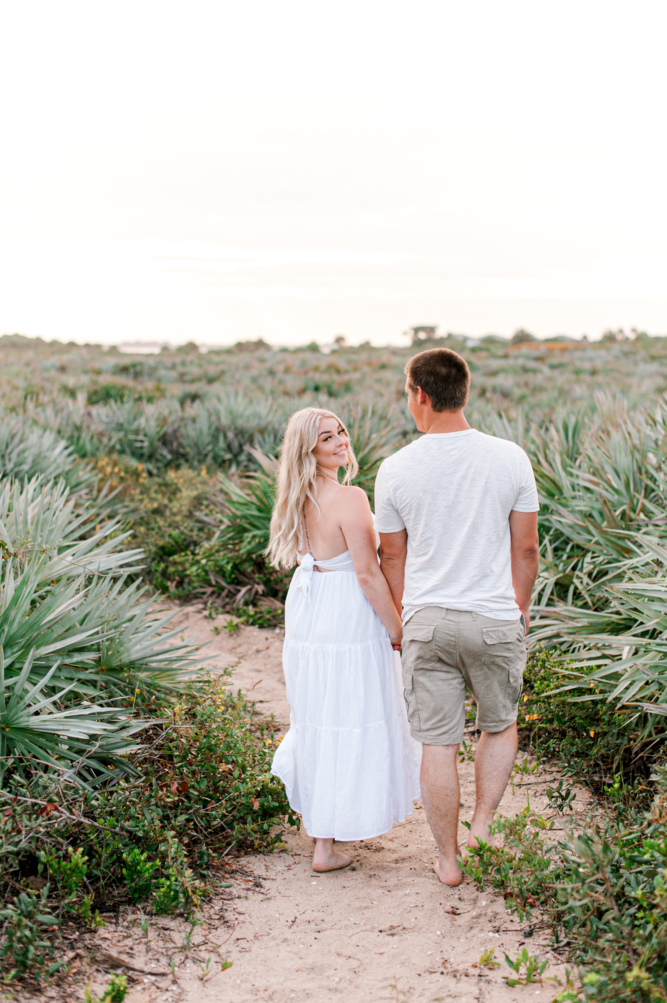Couple walking through a trail near the beach and girl looks back and smiles at the camera during her engagement session Orlando hair salons