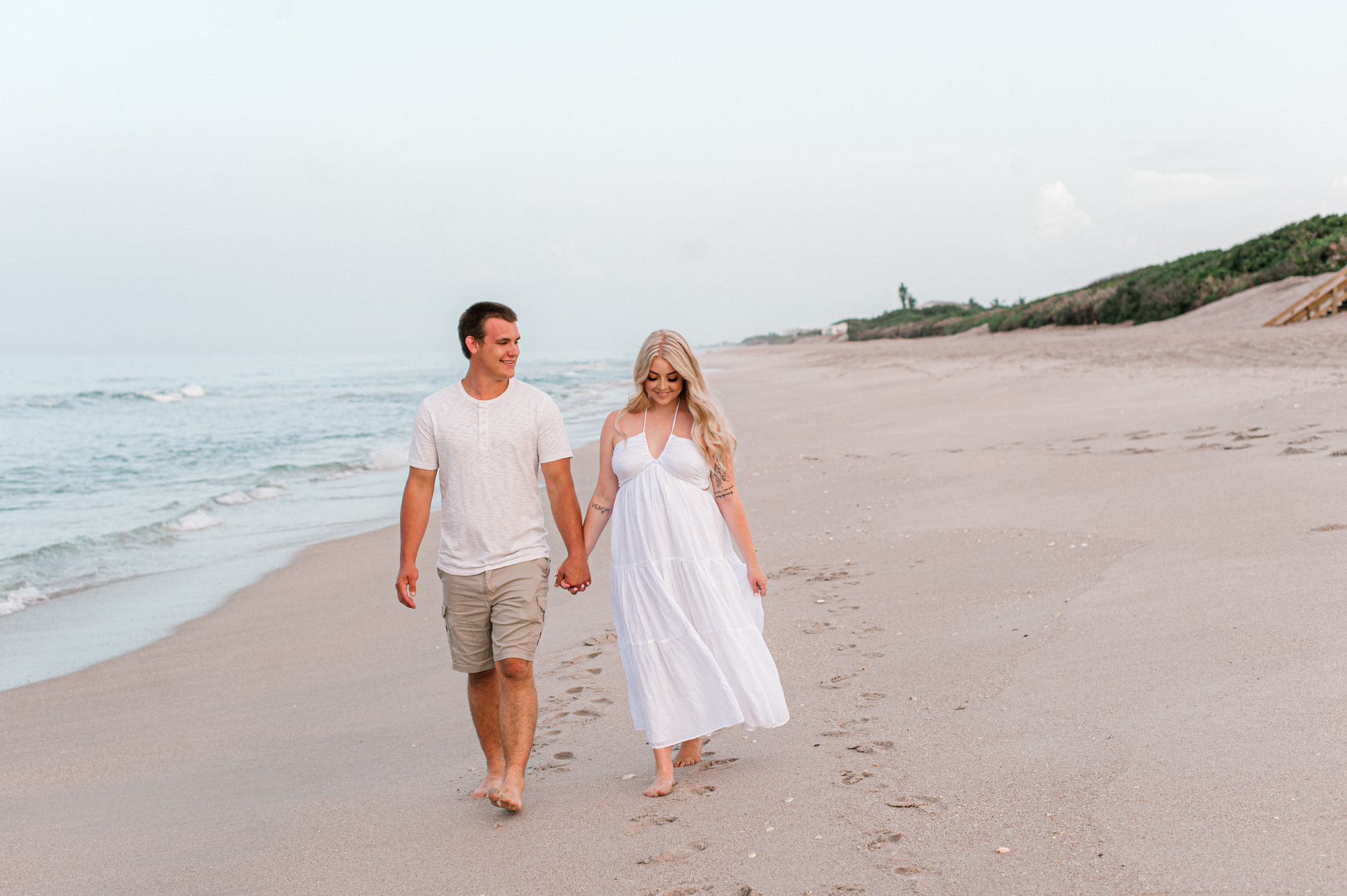 Newly engaged couple walking along the beach during their engagement session wearing neutral colors and holding hands.