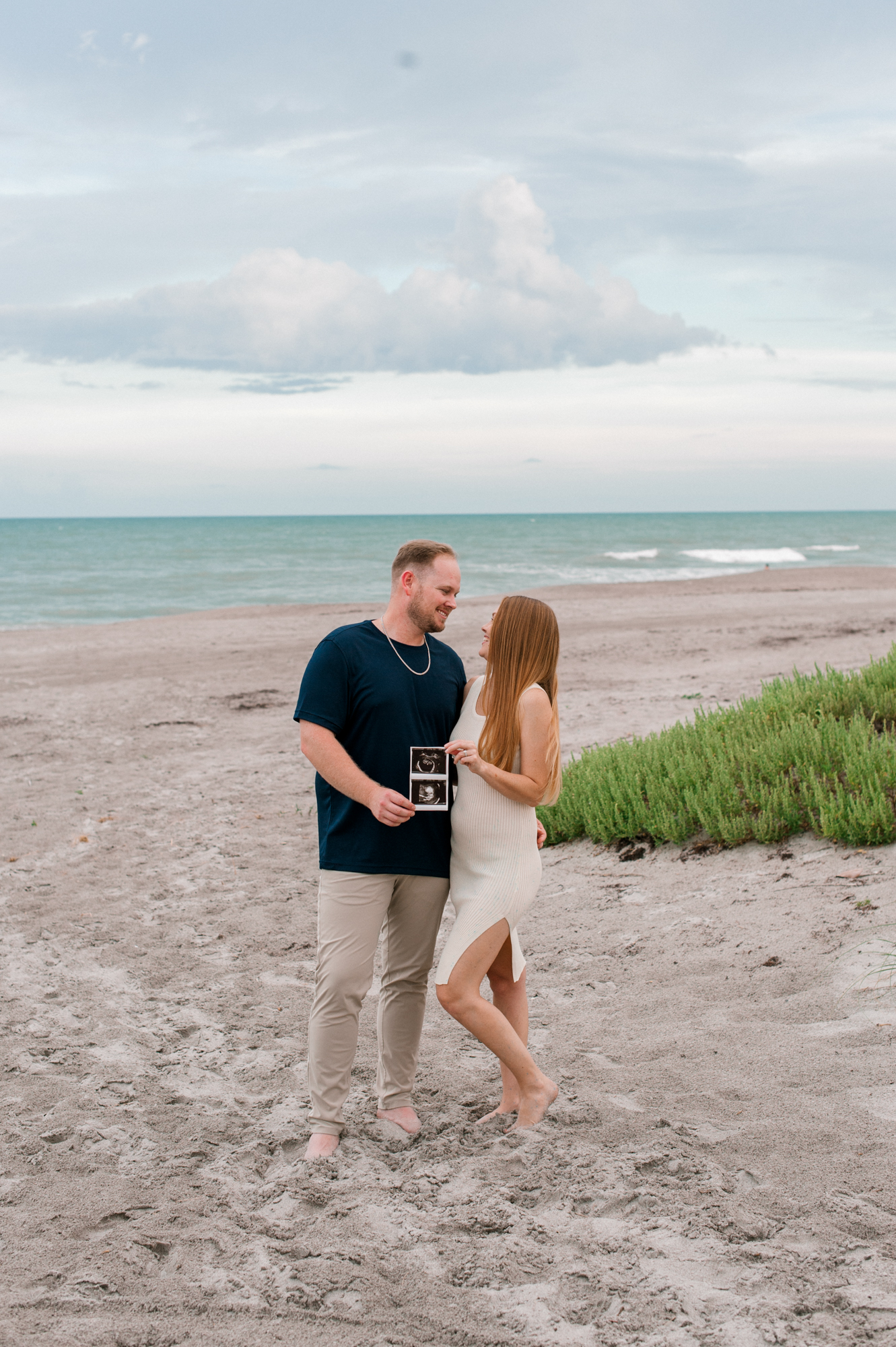 New parents pose on the beach near the dunes holding an ultrasound picture of their growing baby boy orlando prenatal massage