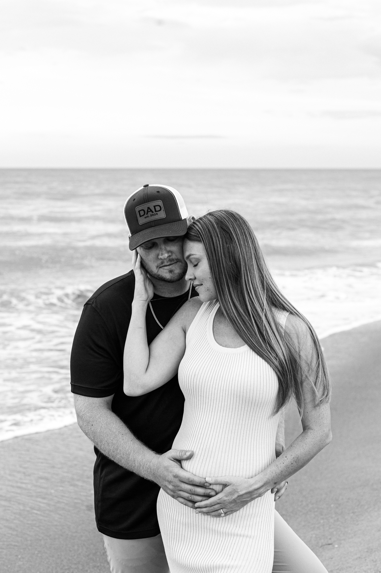 New parents stand near the ocean holding the moms pregnant belly while snuggled close together orlando prenatal massage
