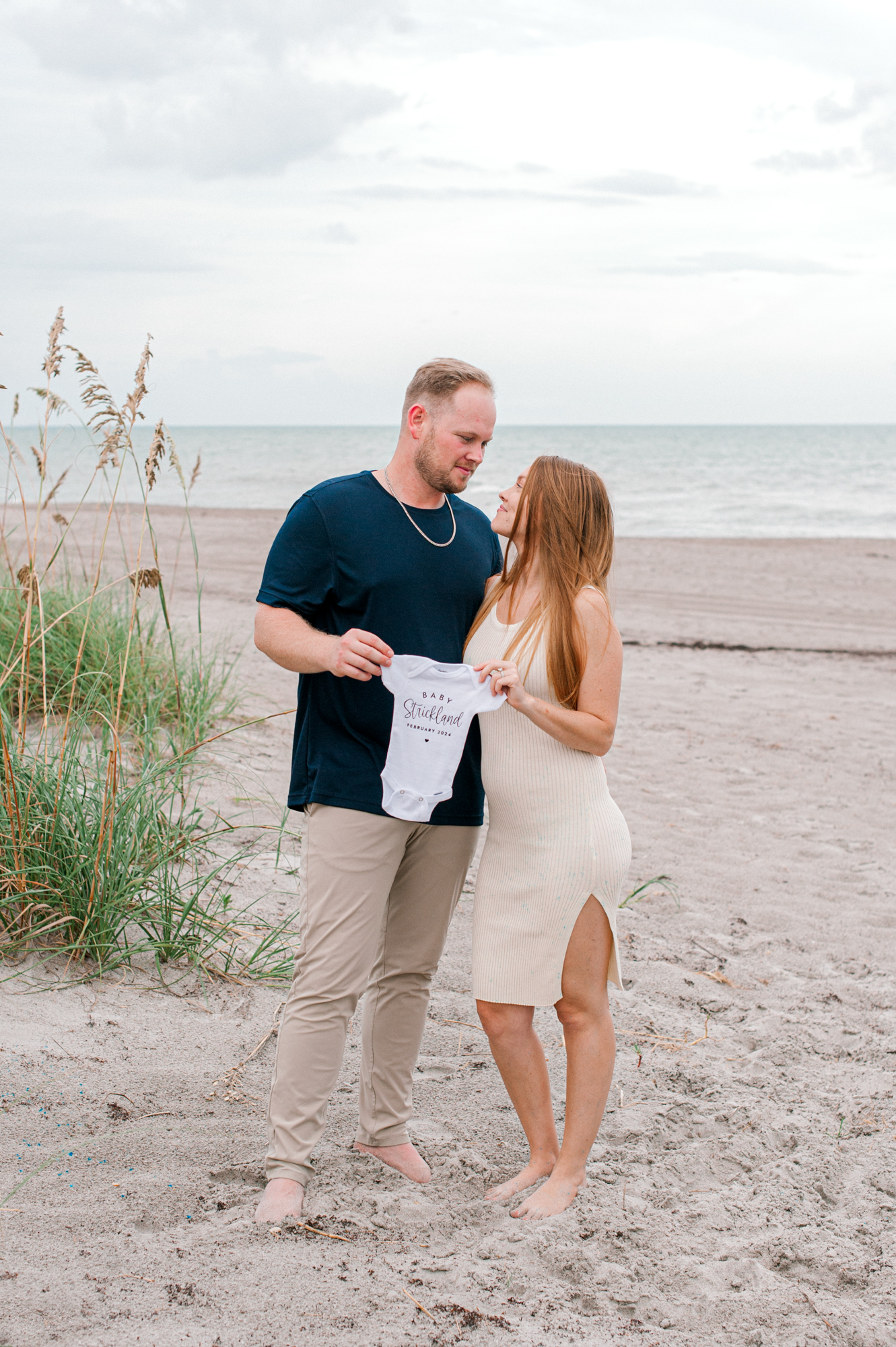 New parents stand near the dunes holding a baby onesie so they can announce to friends and family their exciting news. Orlando Prenatal Massage