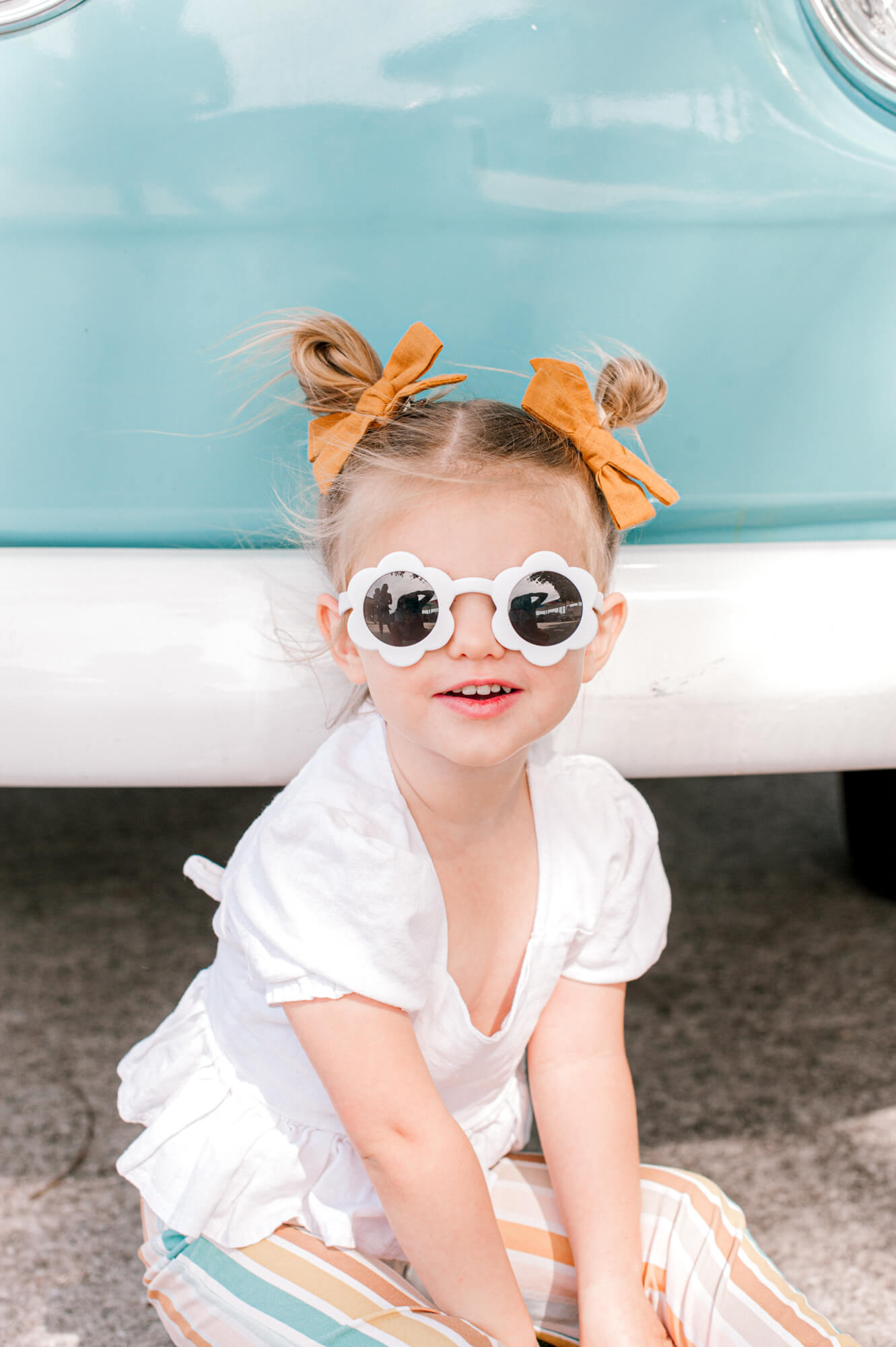 Adorable little girl in flower sunnies with space buns and the cutest outfit sitting in front of a blue VW bus