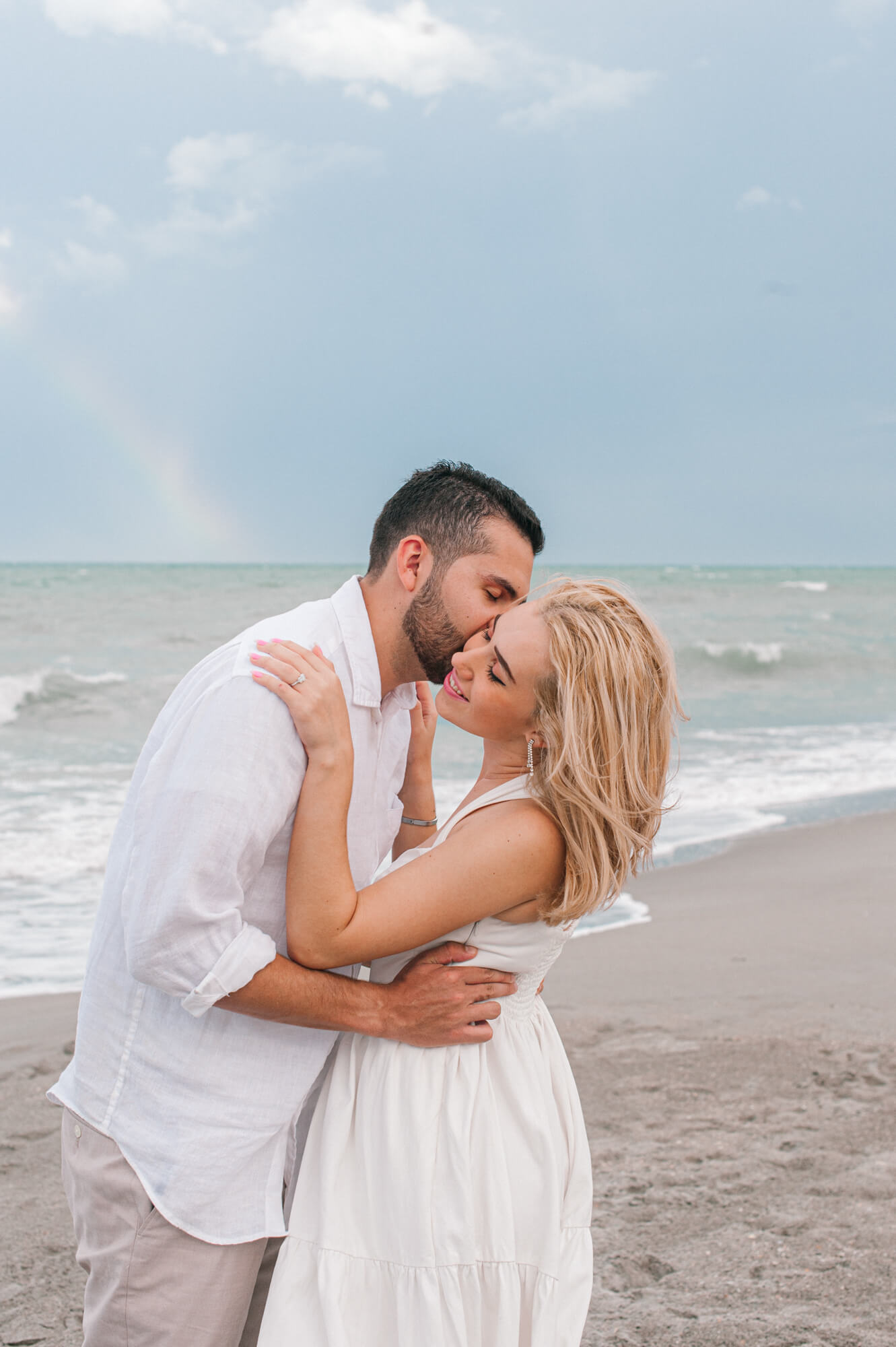 Man kissing his fiance on the cheek near the water line of the beach with a rainbow in the background over the water