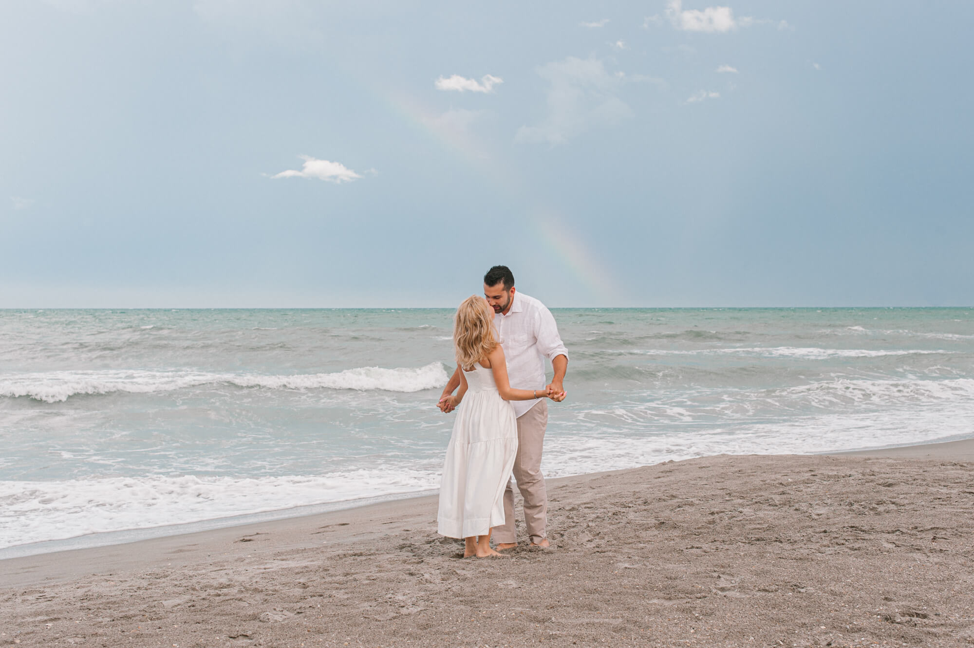 Couple dancing on the beach near the water line in all white with a beautiful rainbow in the background spas in Daytona Beach