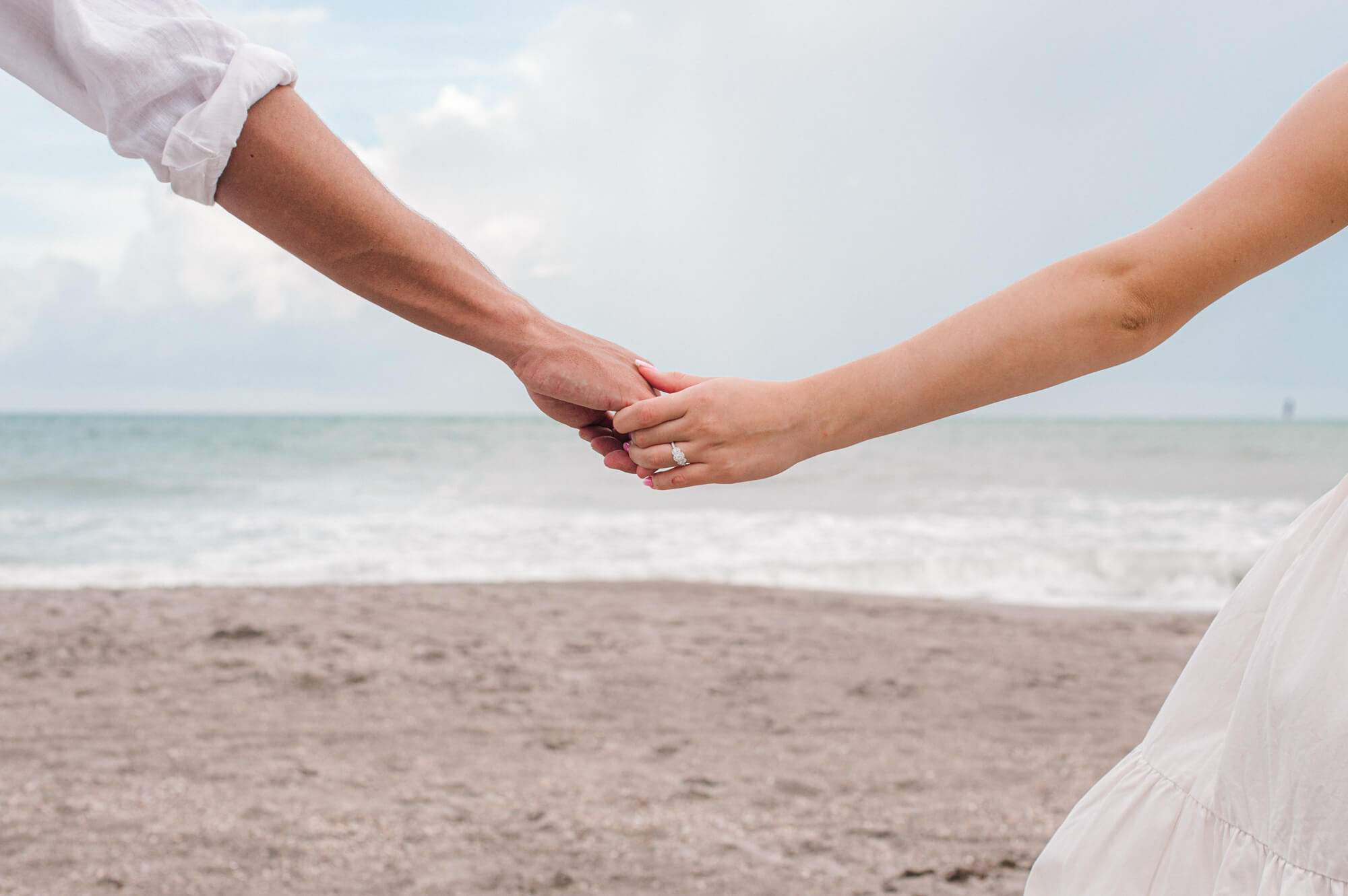 couple holding hands facing the ocean with ring in focus