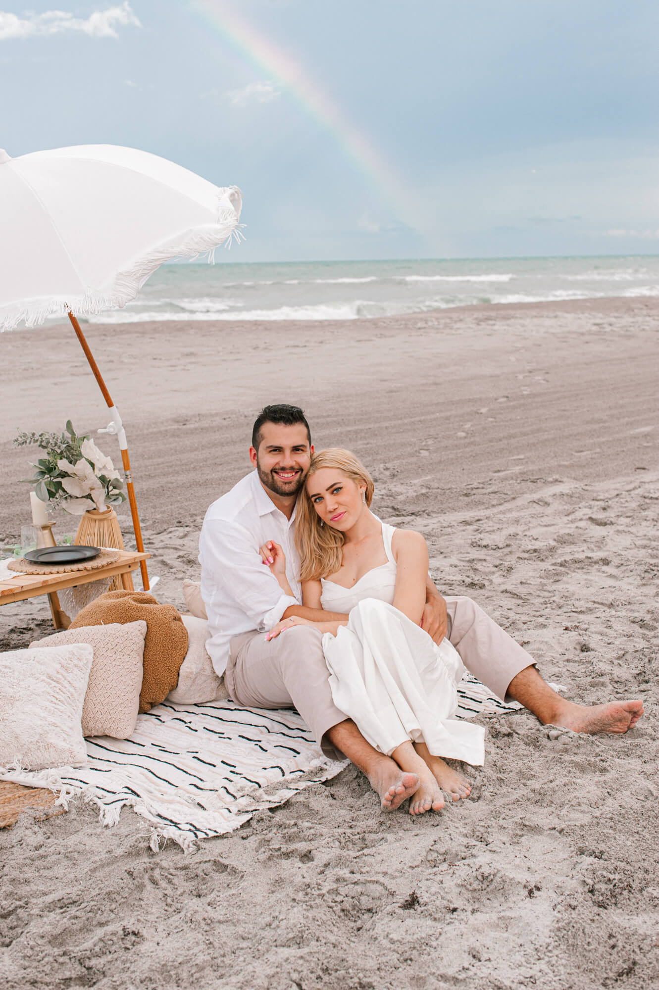 Adorable couple cuddling on the beach during their beach picnic with a rainbow in the background over the water